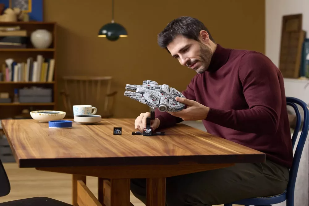 A man holds a Star Wars Millennium Falcon Lego set. He is sitting at a wooden table. Other objects are on the table.