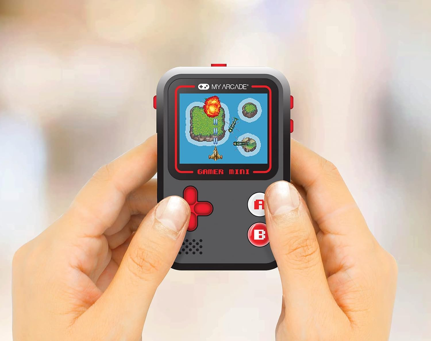 Mains tenant un My Arcade Gamer Mini gris, affichant un jeu pixelisé avec un avion. Boutons et accents rouges. Arrière-plan flou.
