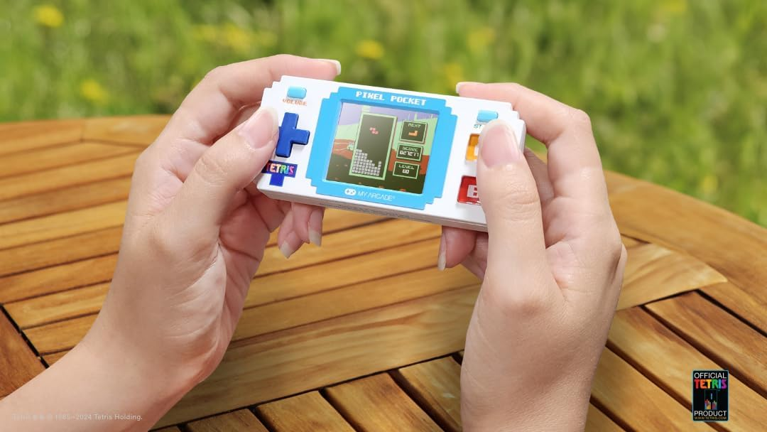 Une personne tient un jeu Tetris Pixel Pocket. Le jeu est blanc avec un bord et des boutons bleus. Le fond est vert flou.