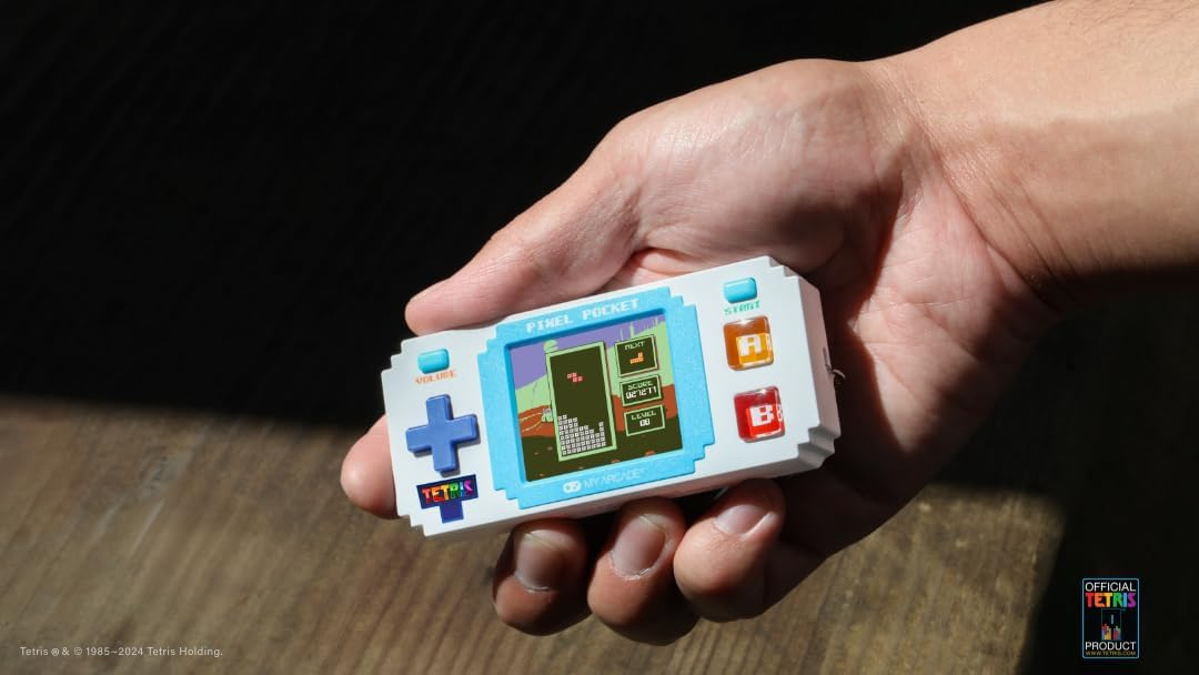 Une main tient un jeu Tetris Pixel Pocket blanc et bleu. L'écran affiche le jeu, avec des blocs colorés. Les boutons sont visibles.