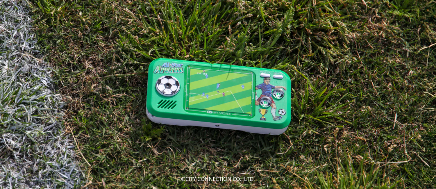 Une console de jeu portable verte sur de l'herbe. L'écran affiche un match de foot. On voit une ligne blanche et de l'herbe.