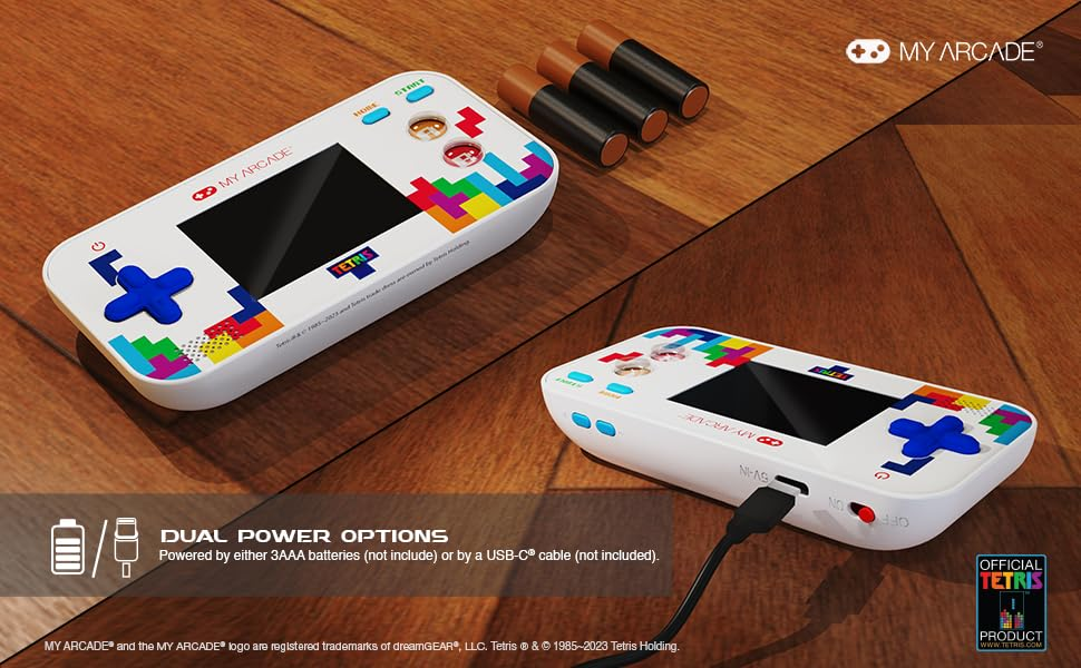 Deux consoles de jeu portables blanches avec des motifs Tetris. L'une est branchée. Trois piles sont sur une surface en bois. Le logo est visible.