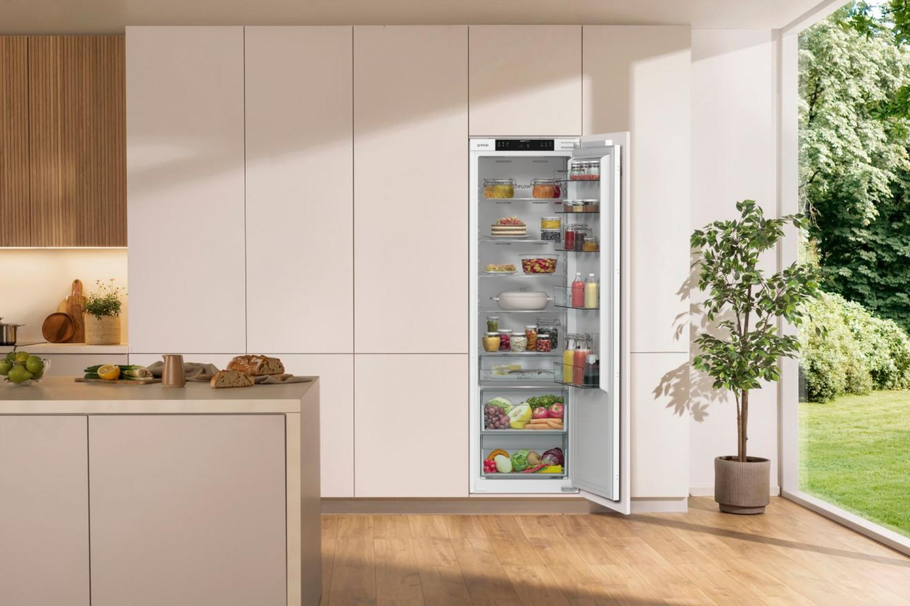 Otwarta lodówka w nowoczesnej kuchni jest wypełniona jedzeniem. Po prawej stronie okno z widokiem na zielony ogród.