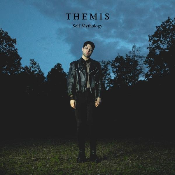 Ein Mann in einer Lederjacke steht auf einem Feld. Bäume bilden einen Hintergrund unter einem blauen Himmel. Text: 'THEMIS' und 'Self Mythology'.