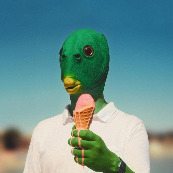 Person mit grüner Maske hält Eiswaffel. Das Eis ist rosa und schmilzt. Der Himmel ist blau.