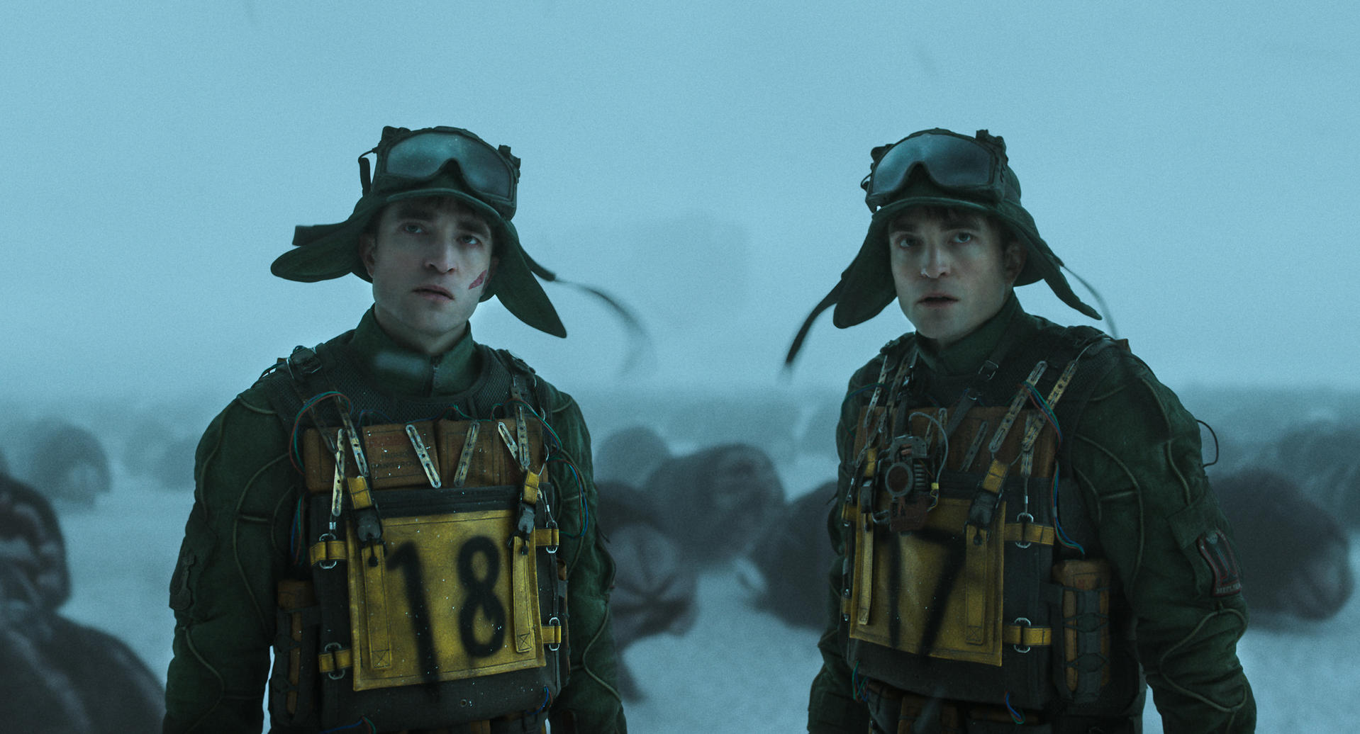 Zwei Männer in grünen Outfits, Hüten und Brillen stehen in einer verschneiten, nebligen Umgebung. Sie tragen Westen mit Nummern.