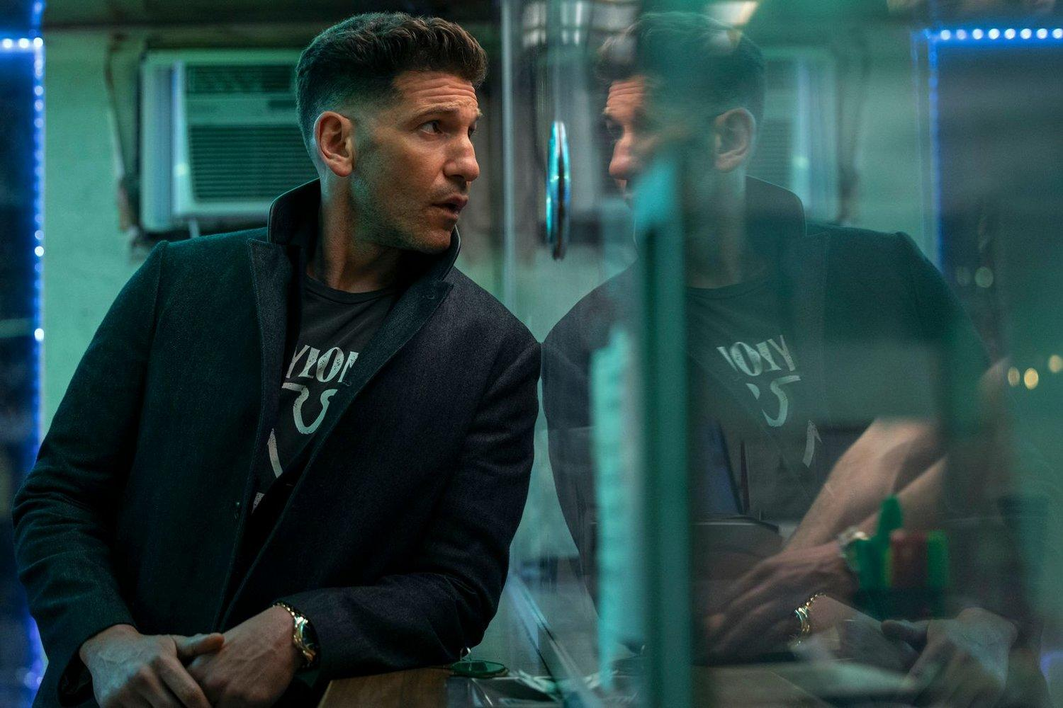 Ein Mann in einem dunklen Mantel betrachtet sein Spiegelbild. Er trägt ein T-Shirt. Der Hintergrund zeigt Lichter und ein Fenster.