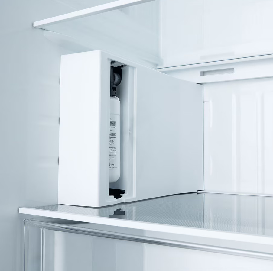 Wewnątrz białej lodówki widoczny jest filtr wody. Wnętrze lodówki jest głównie białe, z półkami i drzwiami.