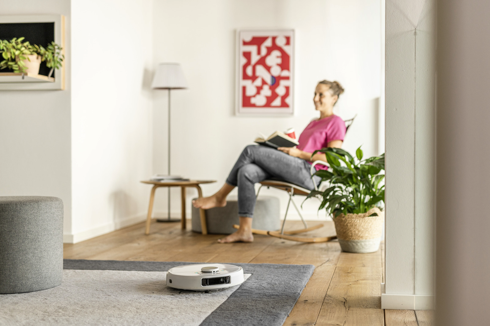 Egy nő egy székben olvas. Egy robotporszívó tisztítja a szőnyeget. Egy növény és egy lámpa látható. A háttér elmosódott.