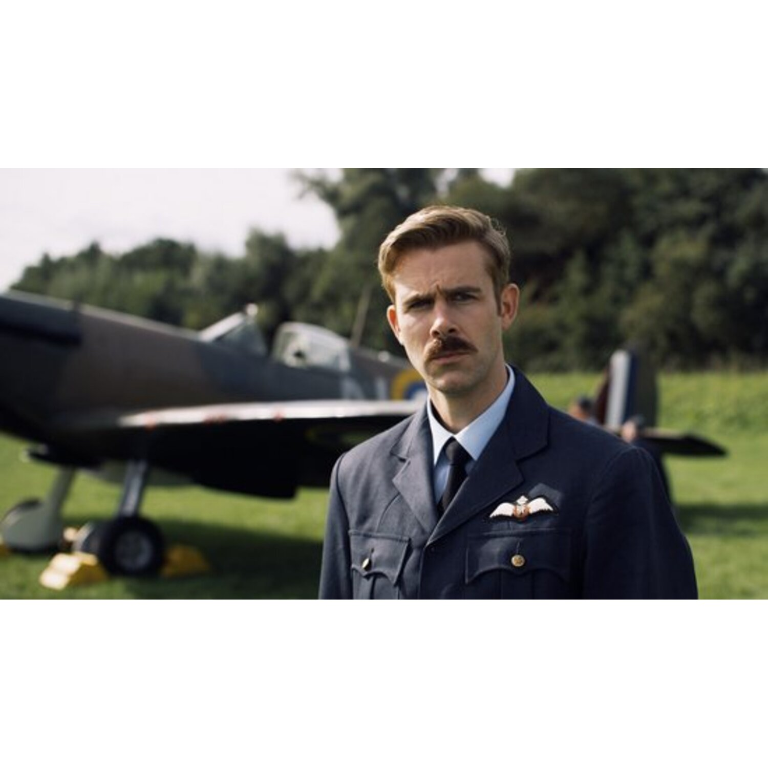 Un homme en uniforme bleu foncé avec une moustache se tient devant un avion. Un champ et des arbres sont en arrière-plan.