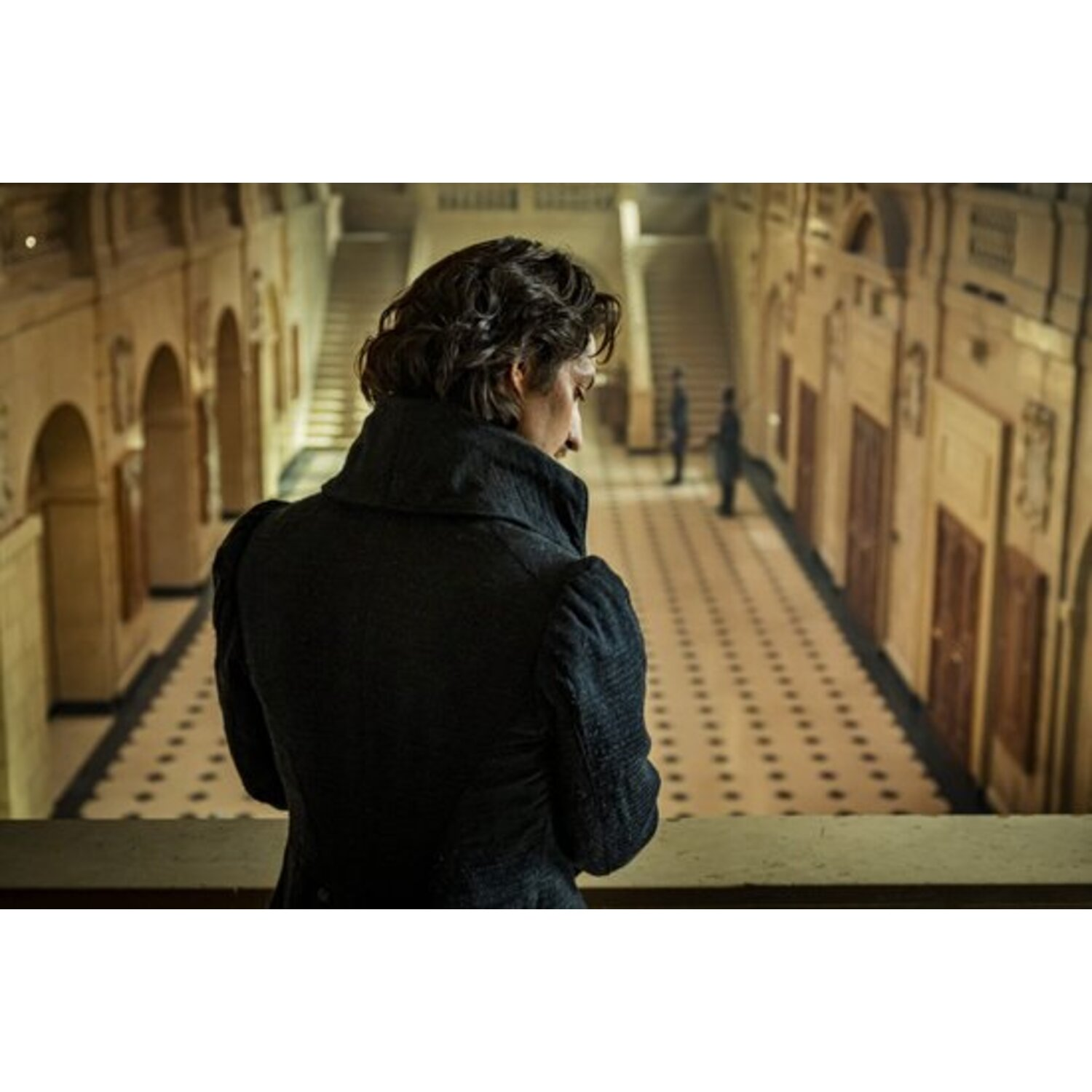 Une personne en manteau noir regarde vers le bas. En arrière-plan, un long couloir avec des portes et des personnages. Le sol est carrelé.
