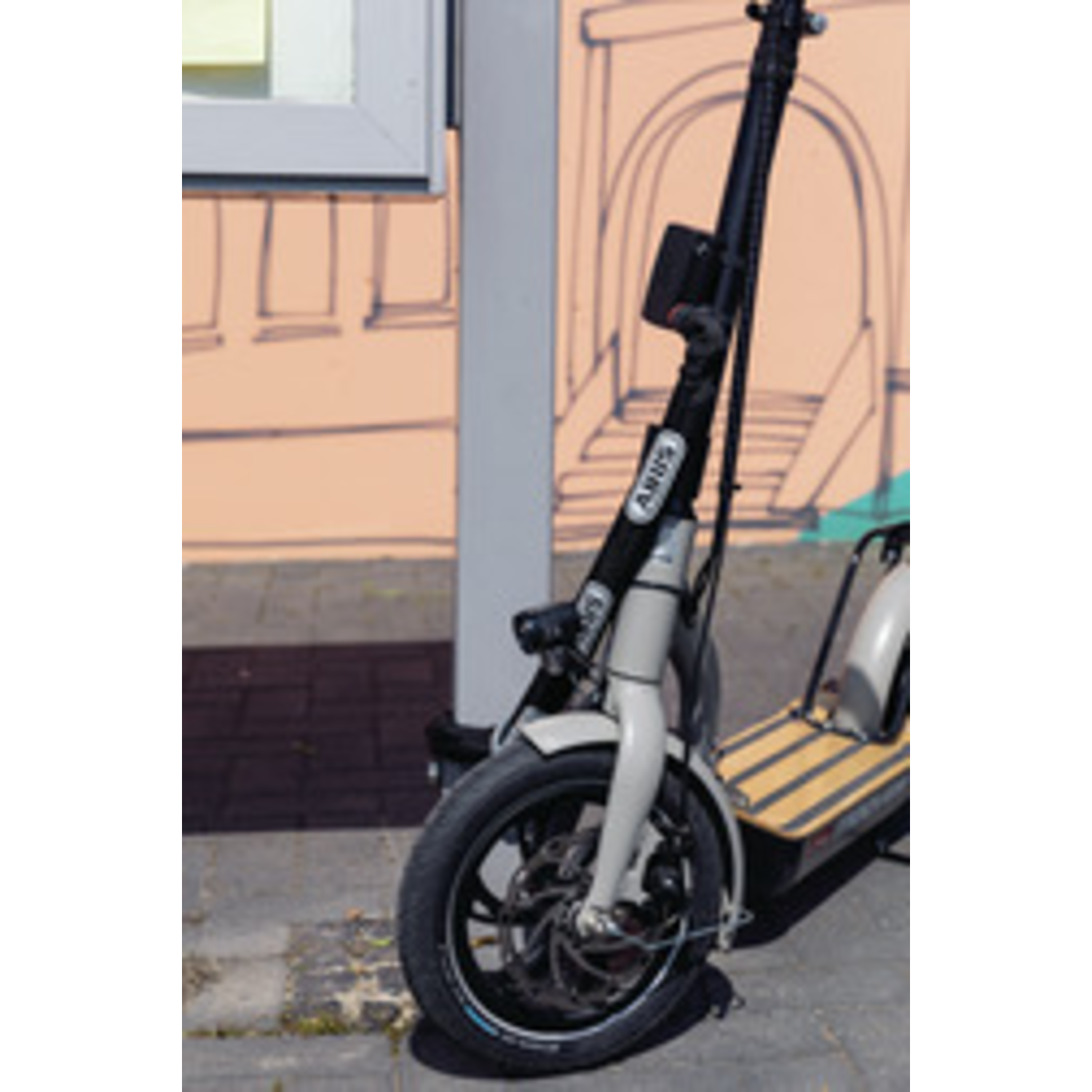 An electric scooter is parked. It has a black tire, white frame, and black handlebar. The background is a pink wall.