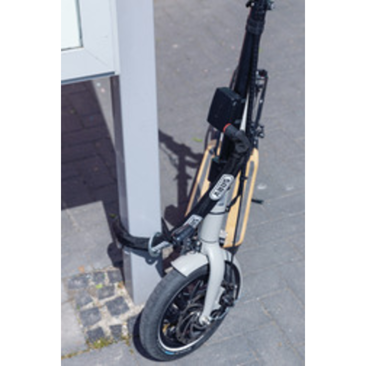 An electric scooter is parked next to a grey pole. The scooter is black and silver with a wooden deck. The background is pavement.