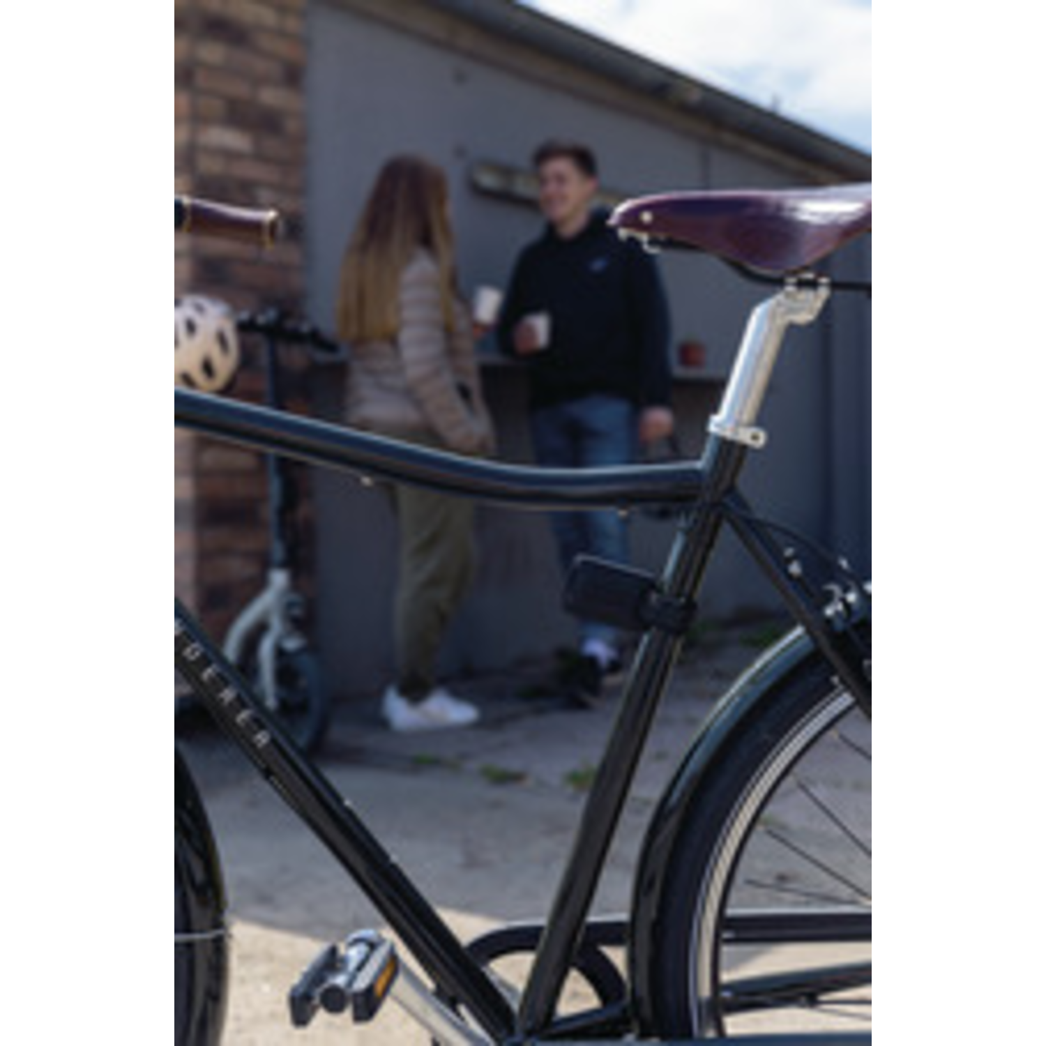 Black bicycle in foreground. Two people blurred in the background, one holding a cup. Scooter visible. Brick wall.