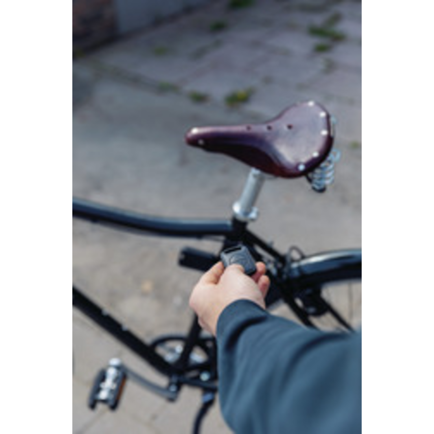 A person holds a black device near a black bicycle. A brown leather seat is visible. The background is blurred with green.