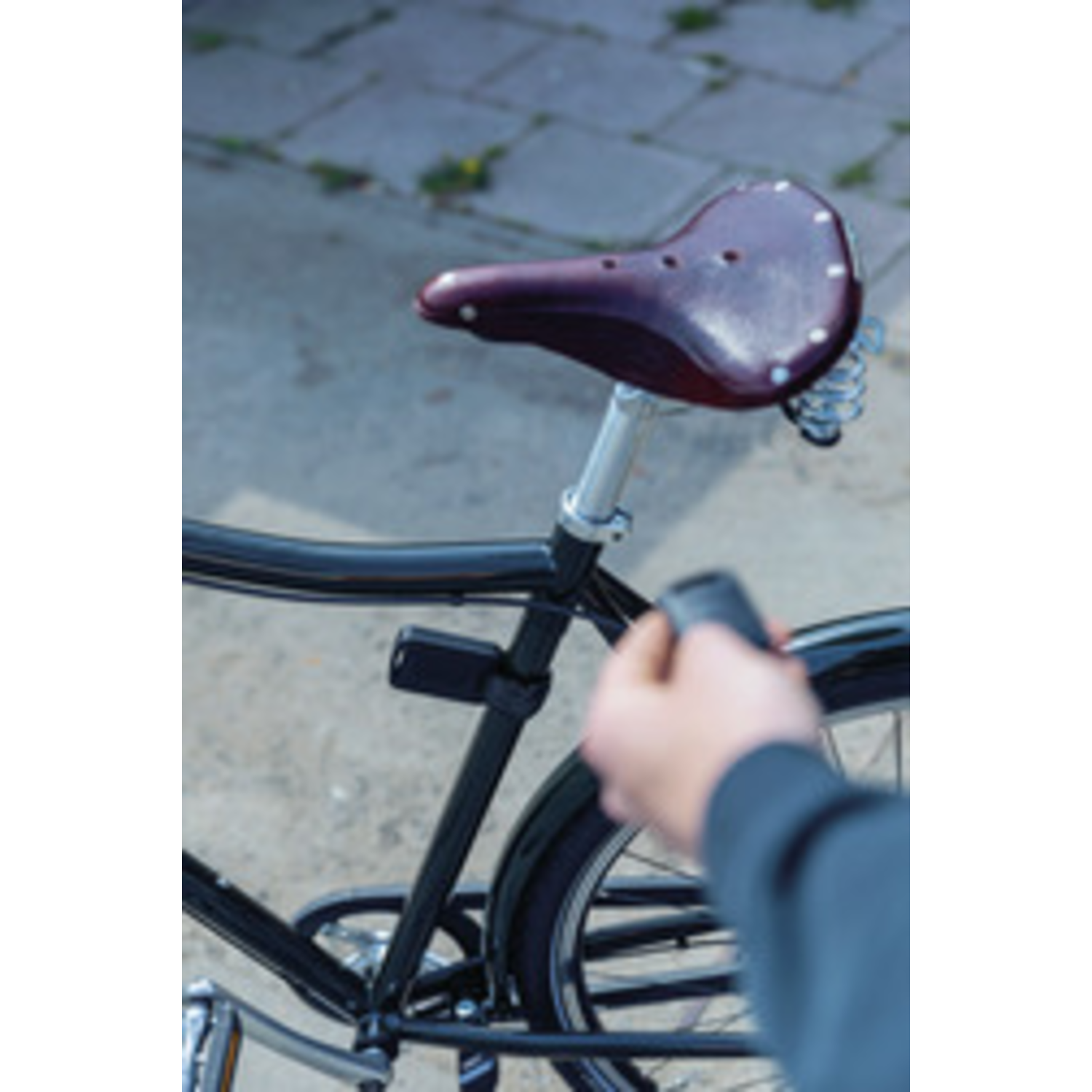 Close-up of a bicycle with a leather saddle. A person's hand is near the rear wheel. The background is a blurry sidewalk.