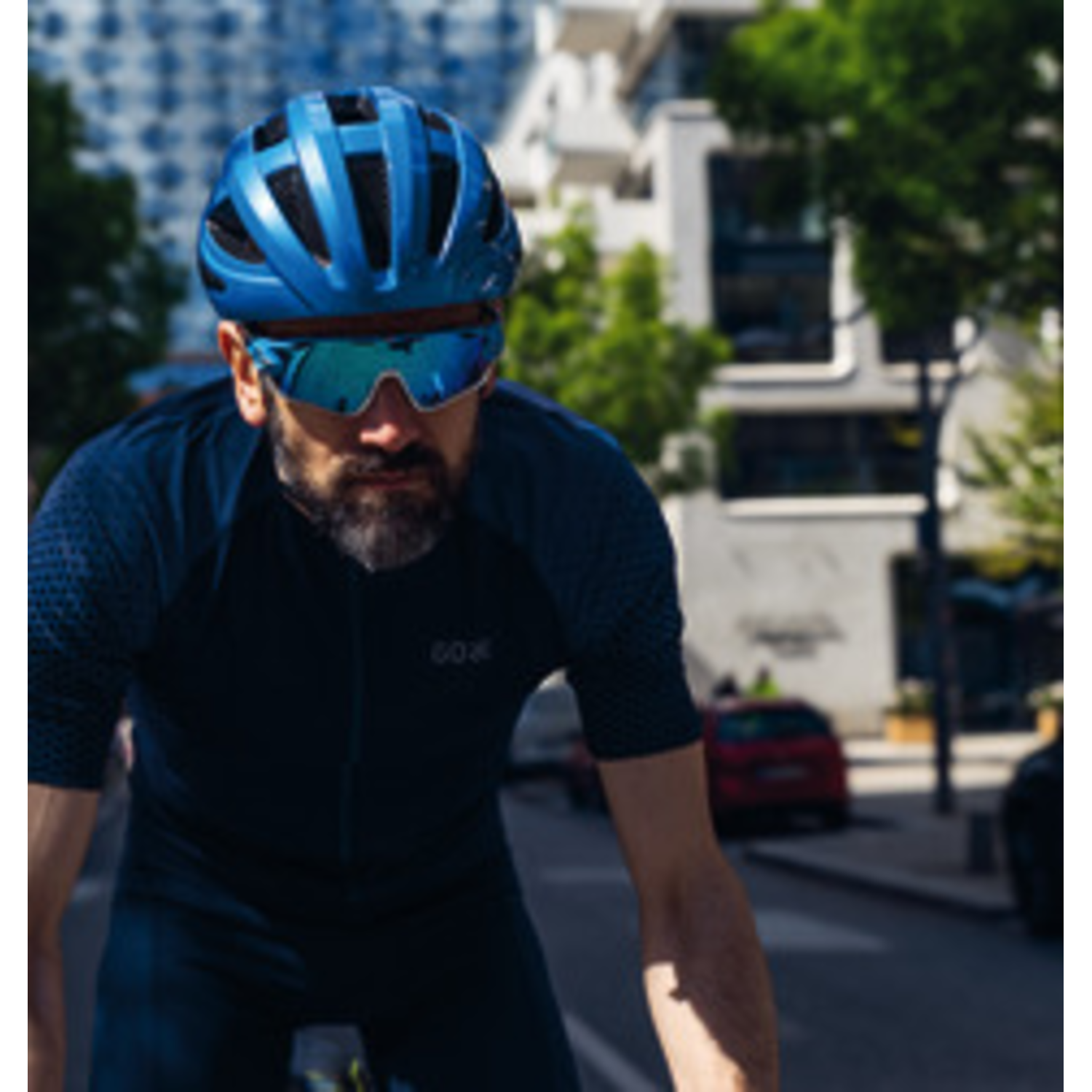 Cyclist in blue helmet and sunglasses on a road. Buildings and trees in the background. Focus on the cyclist.