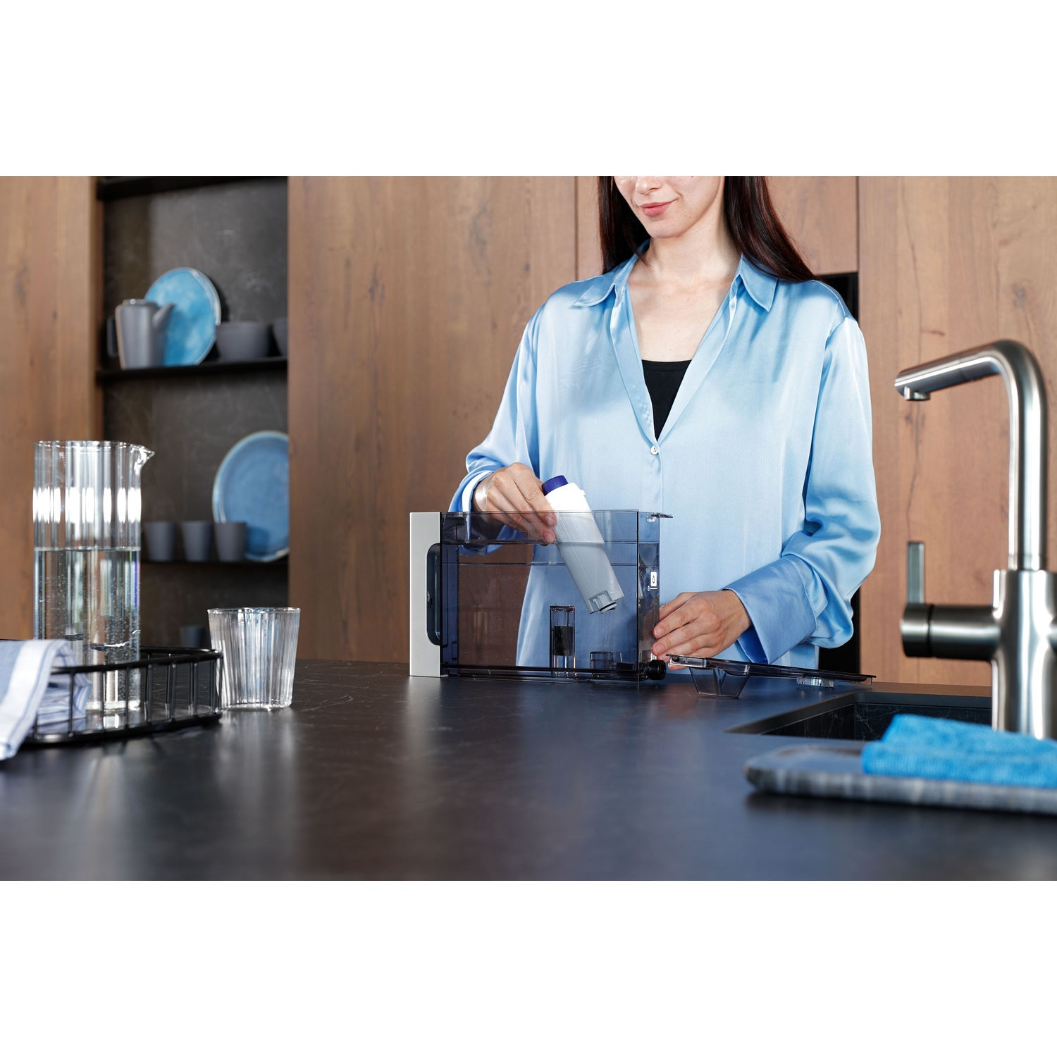 Une femme en chemise bleue insère un filtre dans un récipient d'eau sur un comptoir de cuisine. D'autres articles incluent un verre et un robinet.