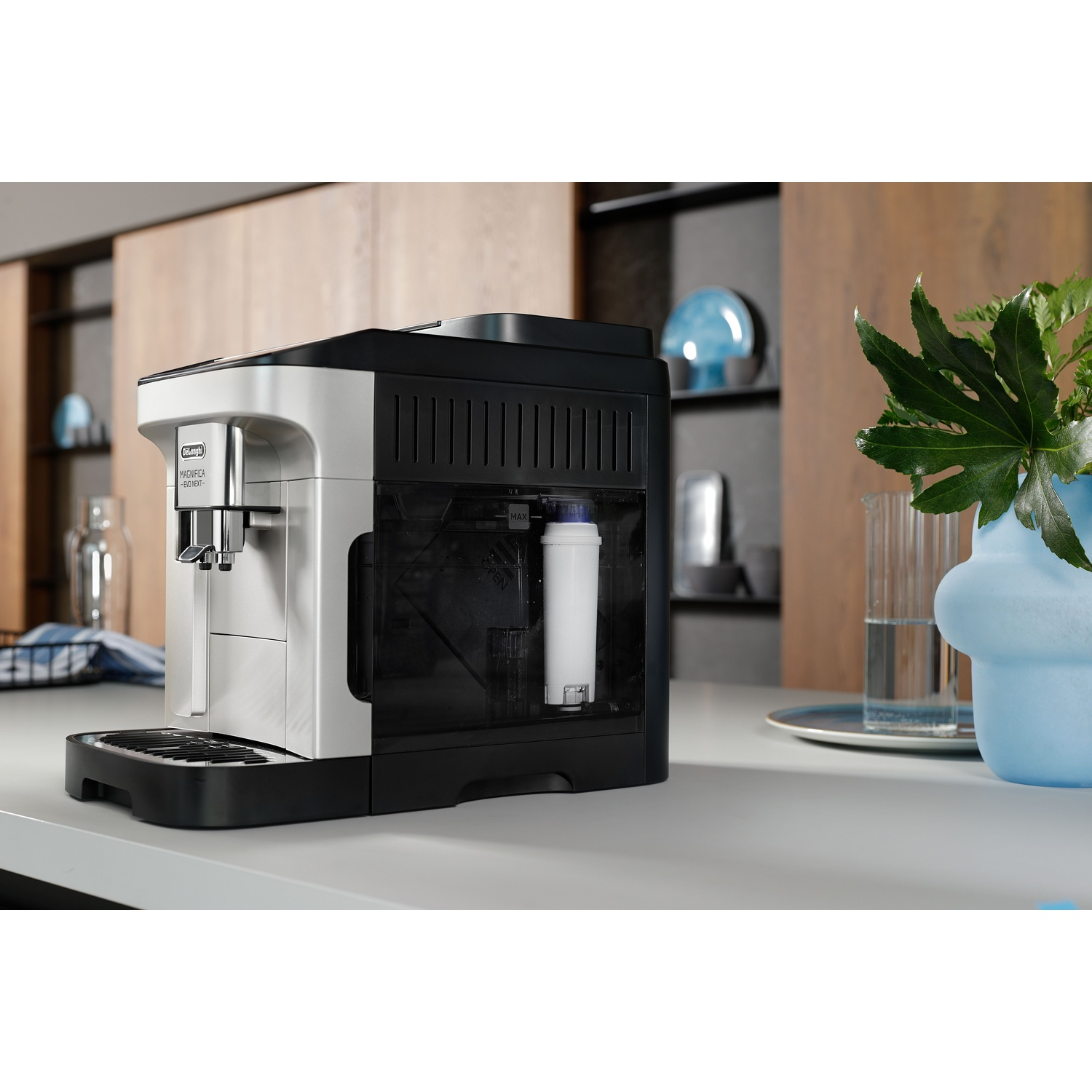 Une machine à expresso De'Longhi sur un comptoir. Un vase bleu avec des feuilles vertes à droite. Des placards en bois en arrière-plan.