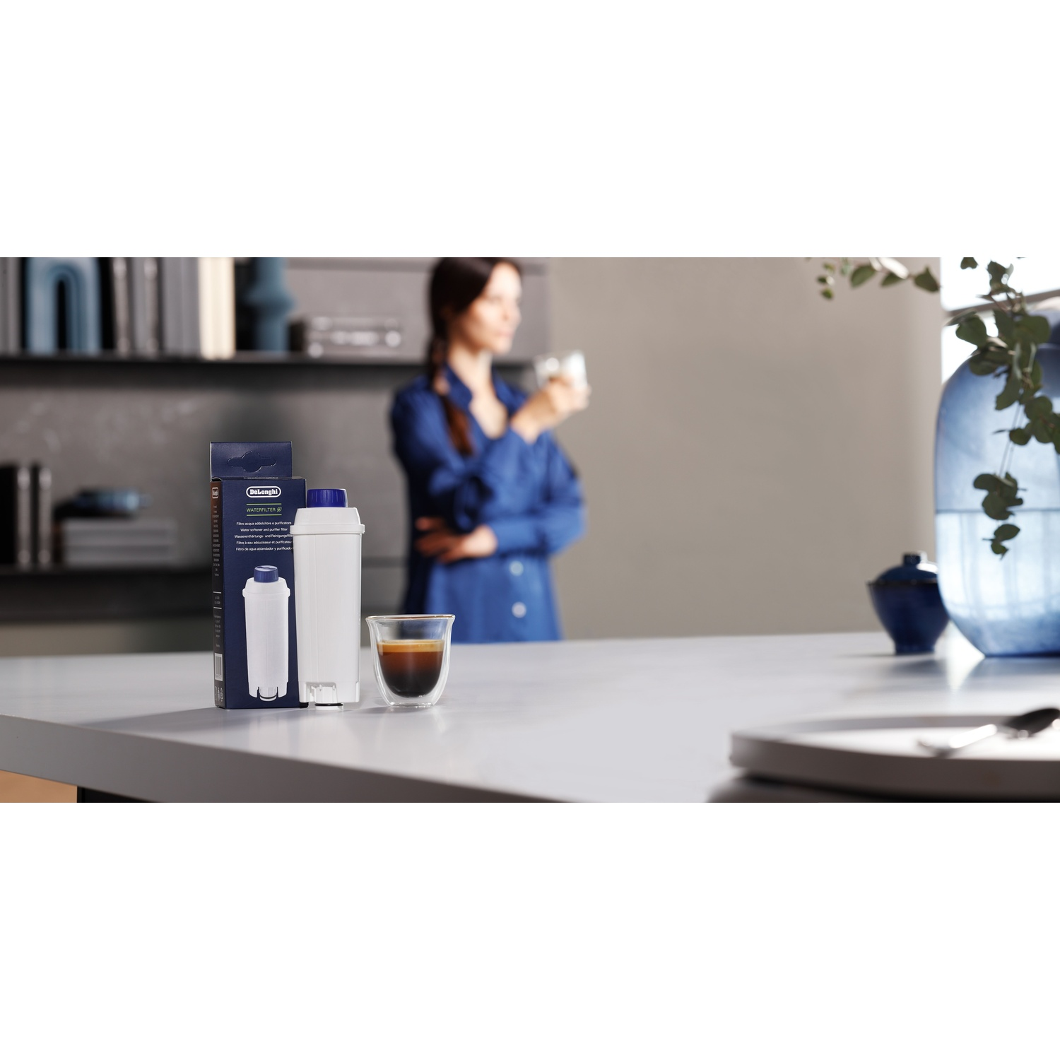 Une préparation de café avec une femme en arrière-plan. Le premier plan comprend un filtre à eau, un verre à café et une boîte. Un vase est également présent.