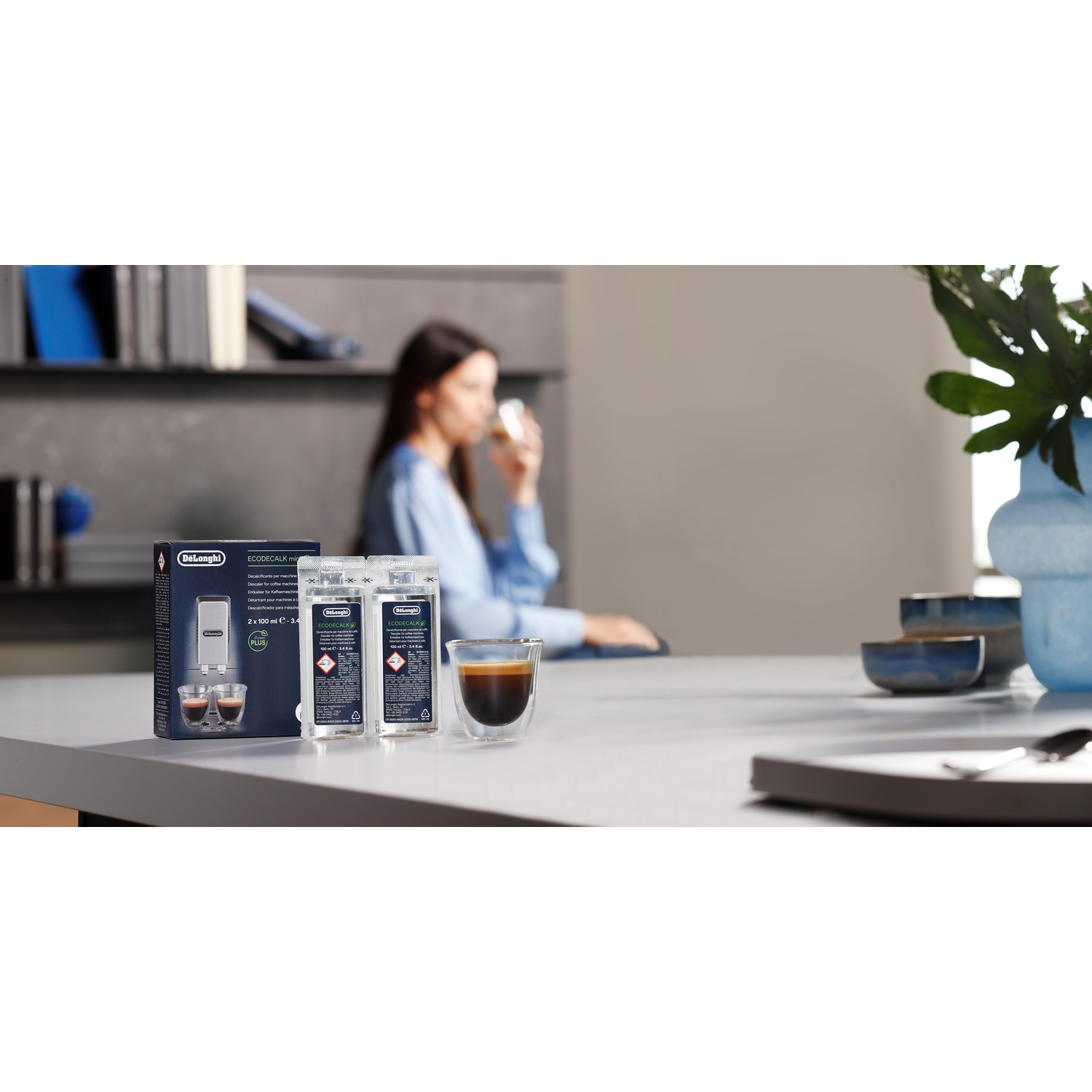 A woman drinks coffee; a box, two bottles, and a coffee cup are on the table. A vase with plants is in the background.