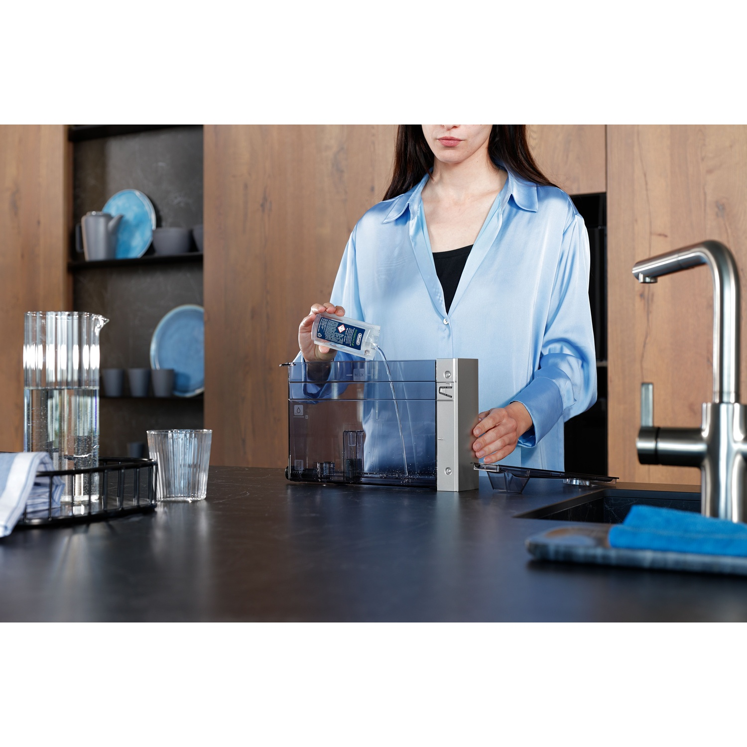 Woman in blue shirt filling a water tank with water. Kitchen setting with glasses, pitcher, and dishes in background.