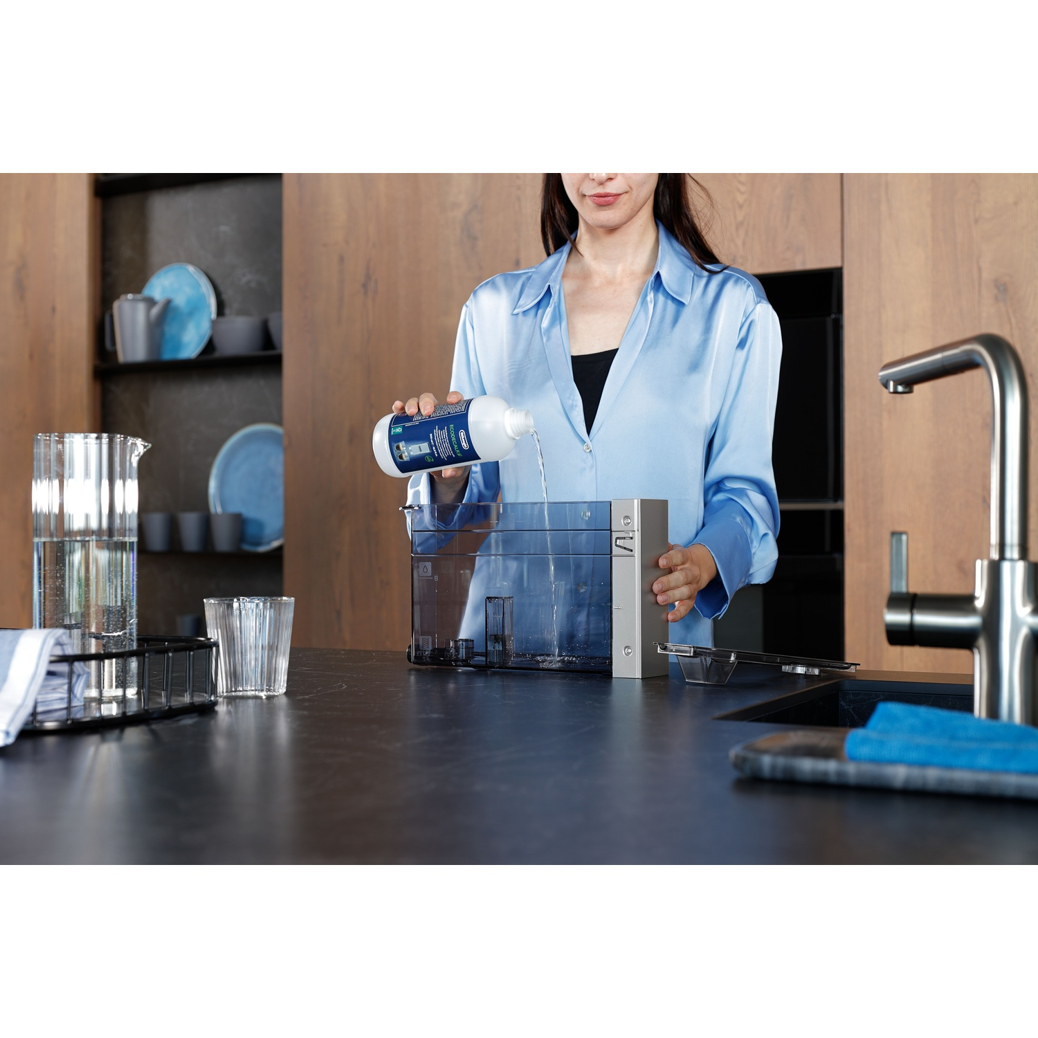 A woman pours liquid from a bottle into a water container. Kitchen setting with a faucet and black countertop. Dishes in background.