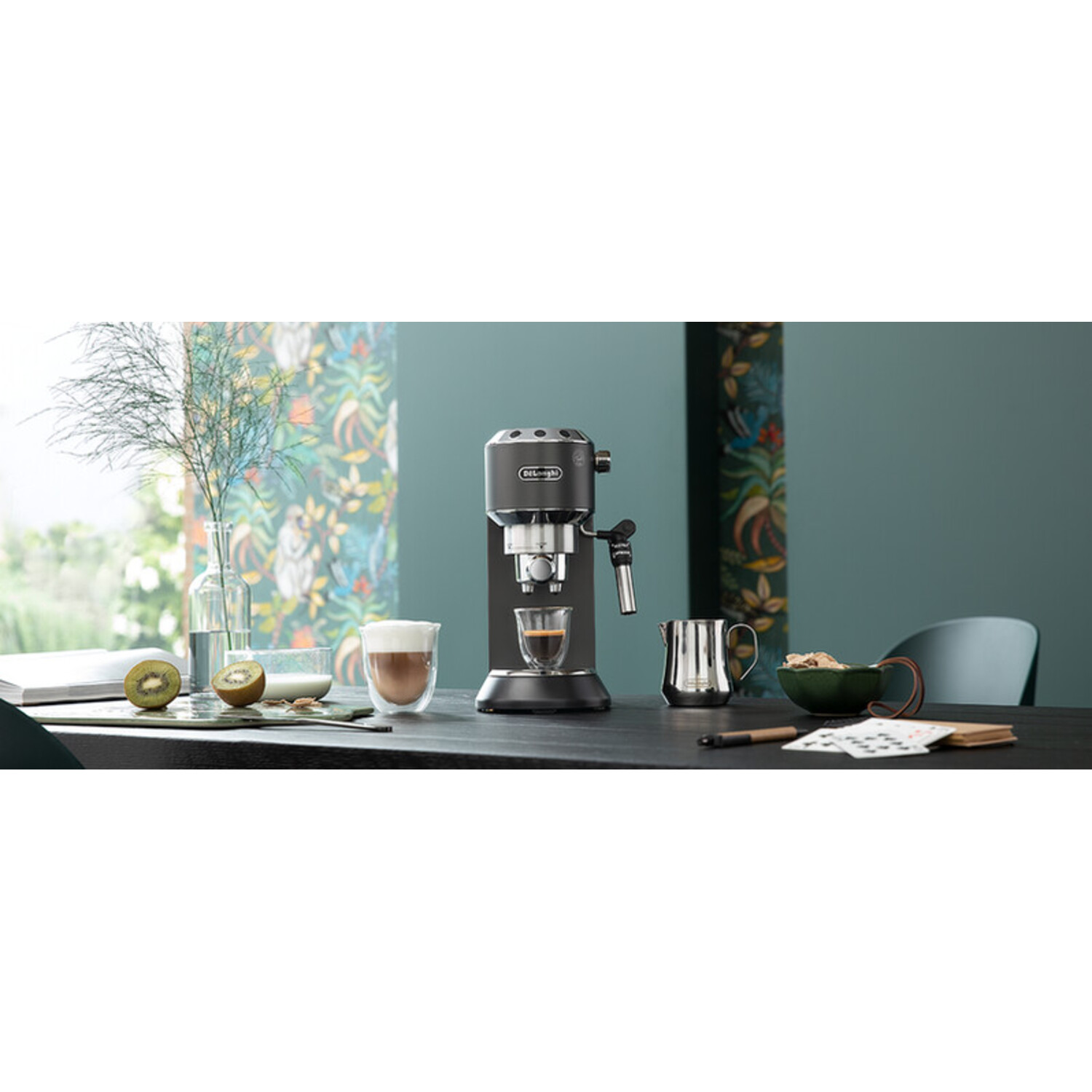 Espressomachine op een tafel met koffie, kiwi en een plant. Achtergrond heeft een bloemenmotief en een blauwgroene muur.