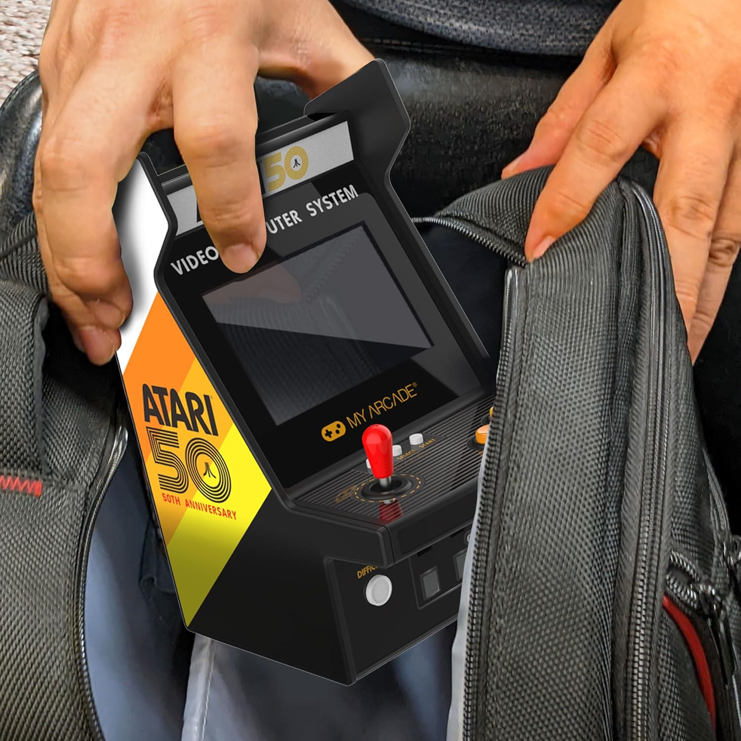 Mains plaçant un jeu d'arcade Atari 50e anniversaire dans un sac noir. Le jeu est noir avec un joystick rouge et des détails jaunes et oranges.