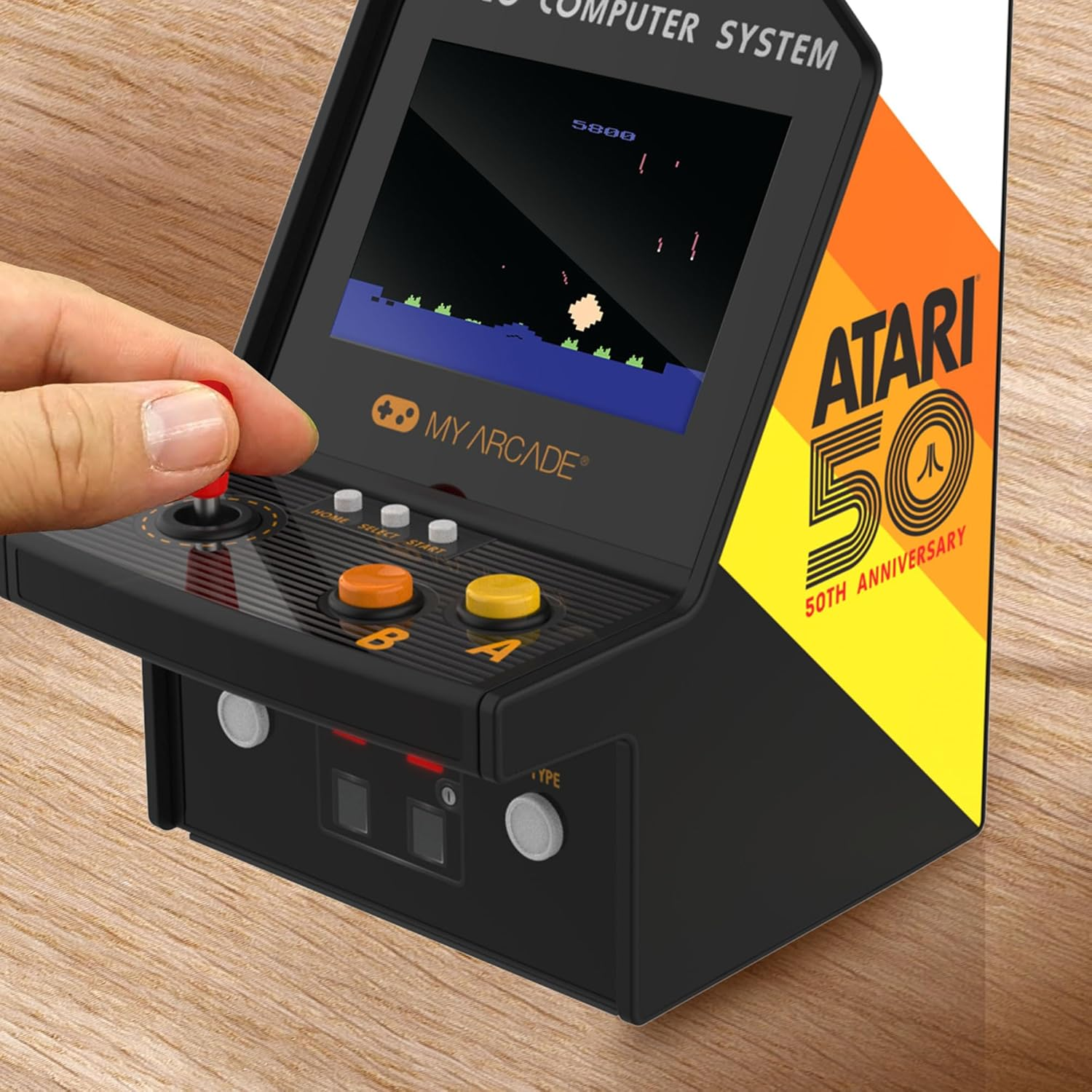 Une main actionne une mini borne d'arcade. L'écran affiche un jeu, et la machine a des boutons et un joystick.