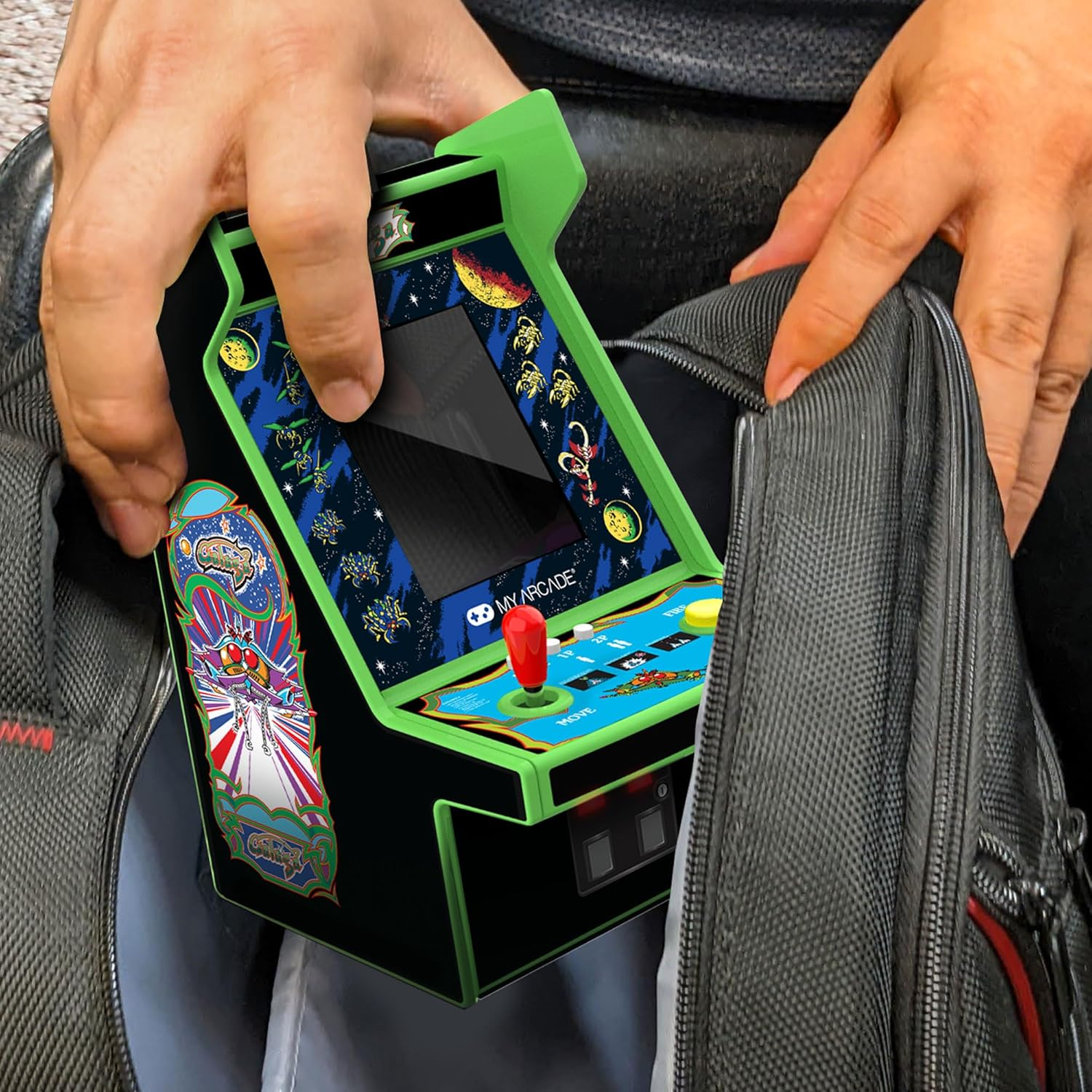 Eine Person hält ein MyArcade Mini-Arcade-Spiel, schwarz und grün. Das Spiel wird in eine graue Tasche gesteckt. Ein roter Joystick ist sichtbar.