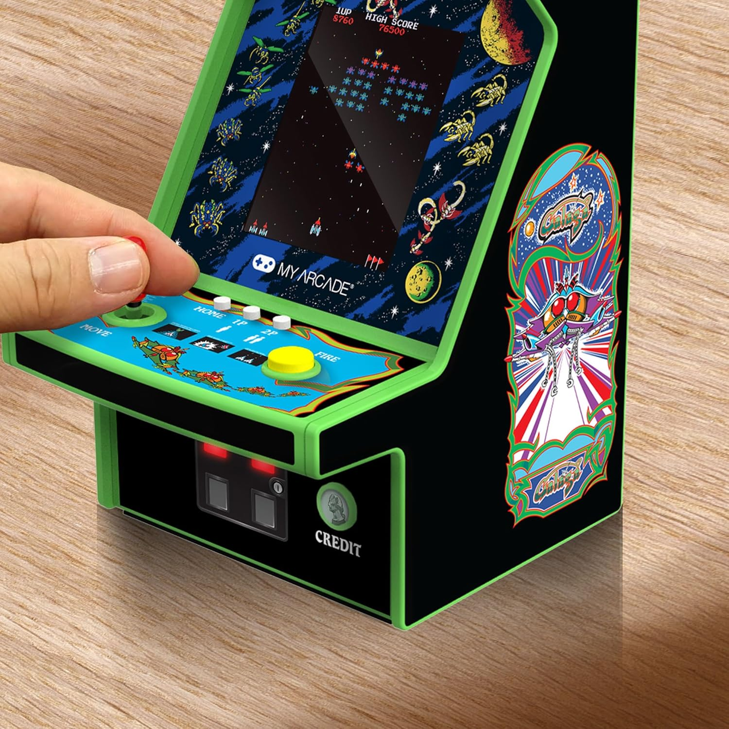 Eine Hand bedient ein Mini-Arcade-Spiel. Das Spiel zeigt ein Space-Invaders-Spiel auf einem schwarz-grünen Automaten auf einer Holzoberfläche.