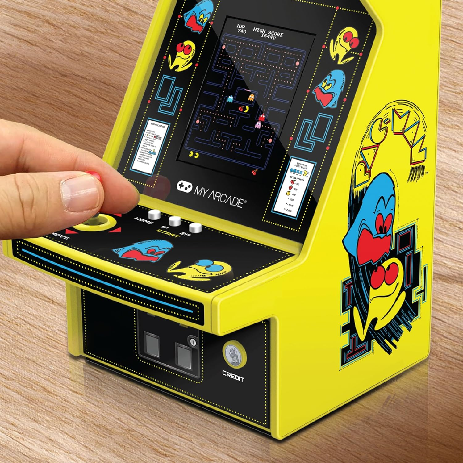 Une main tient un joystick rouge sur une borne d'arcade Pac-Man jaune. L'écran affiche le jeu. Le fond est en bois.