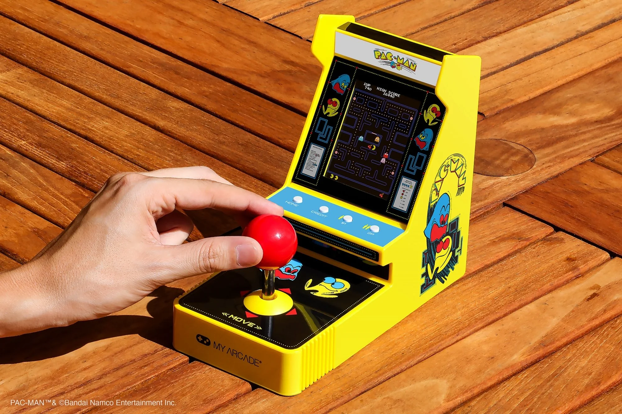 Une main tient une borne d'arcade Pac-Man. Le jeu est jaune avec un joystick rouge, sur une table en bois.