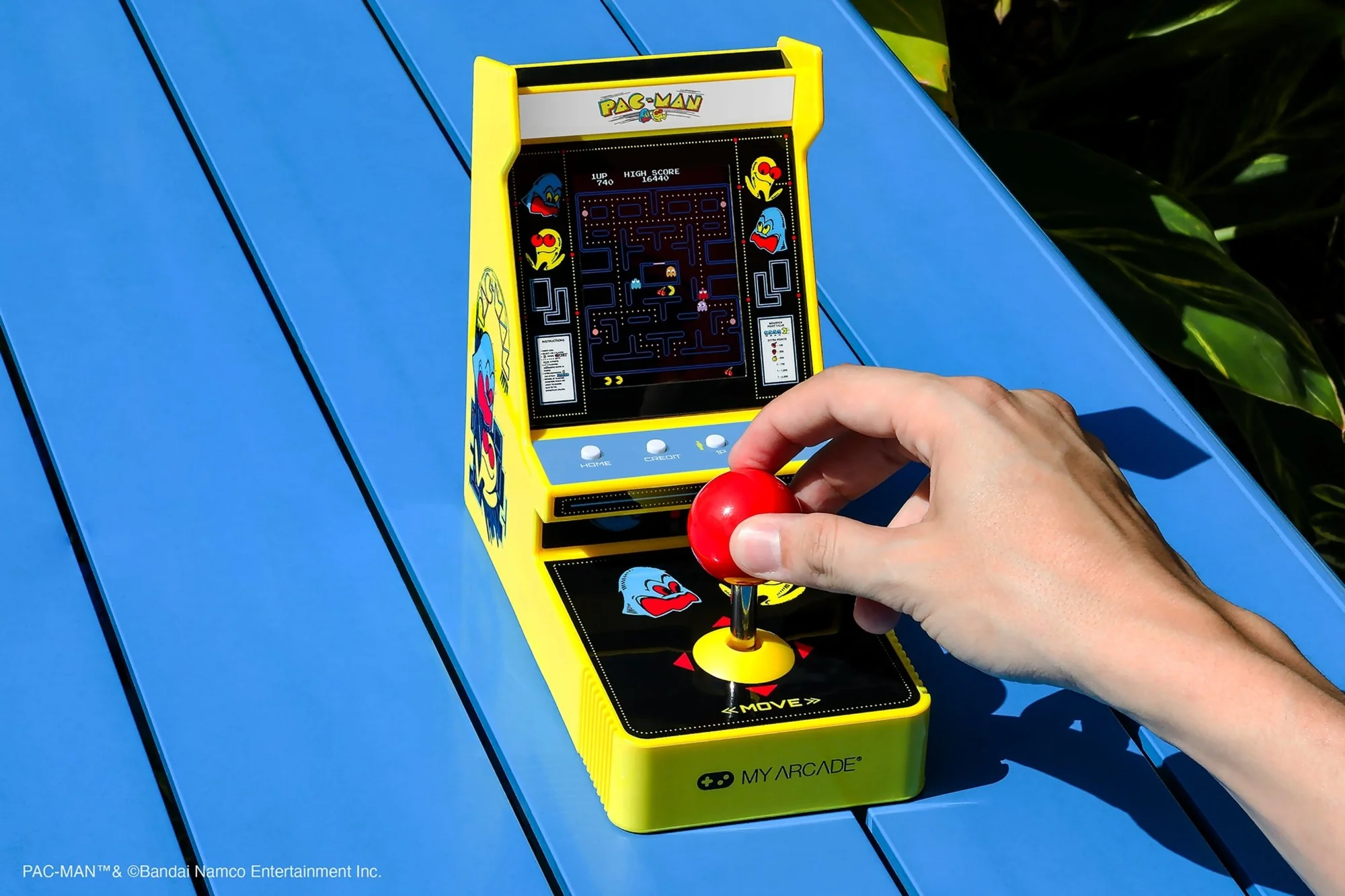 Une borne d'arcade Pac-Man jaune est posée sur une table bleue. Une main tient le joystick rouge. L'écran affiche le jeu.