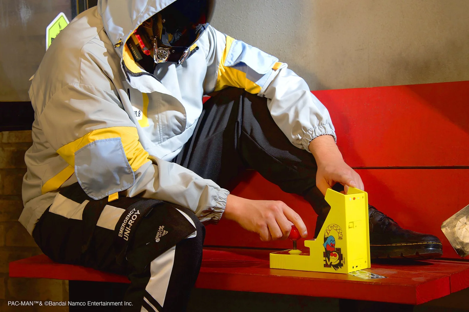 Une personne assise sur un banc rouge tient un jeu d'arcade Pac-Man jaune. Elle porte une veste, un pantalon noir et des chaussures noires. L'arrière-plan est flou.
