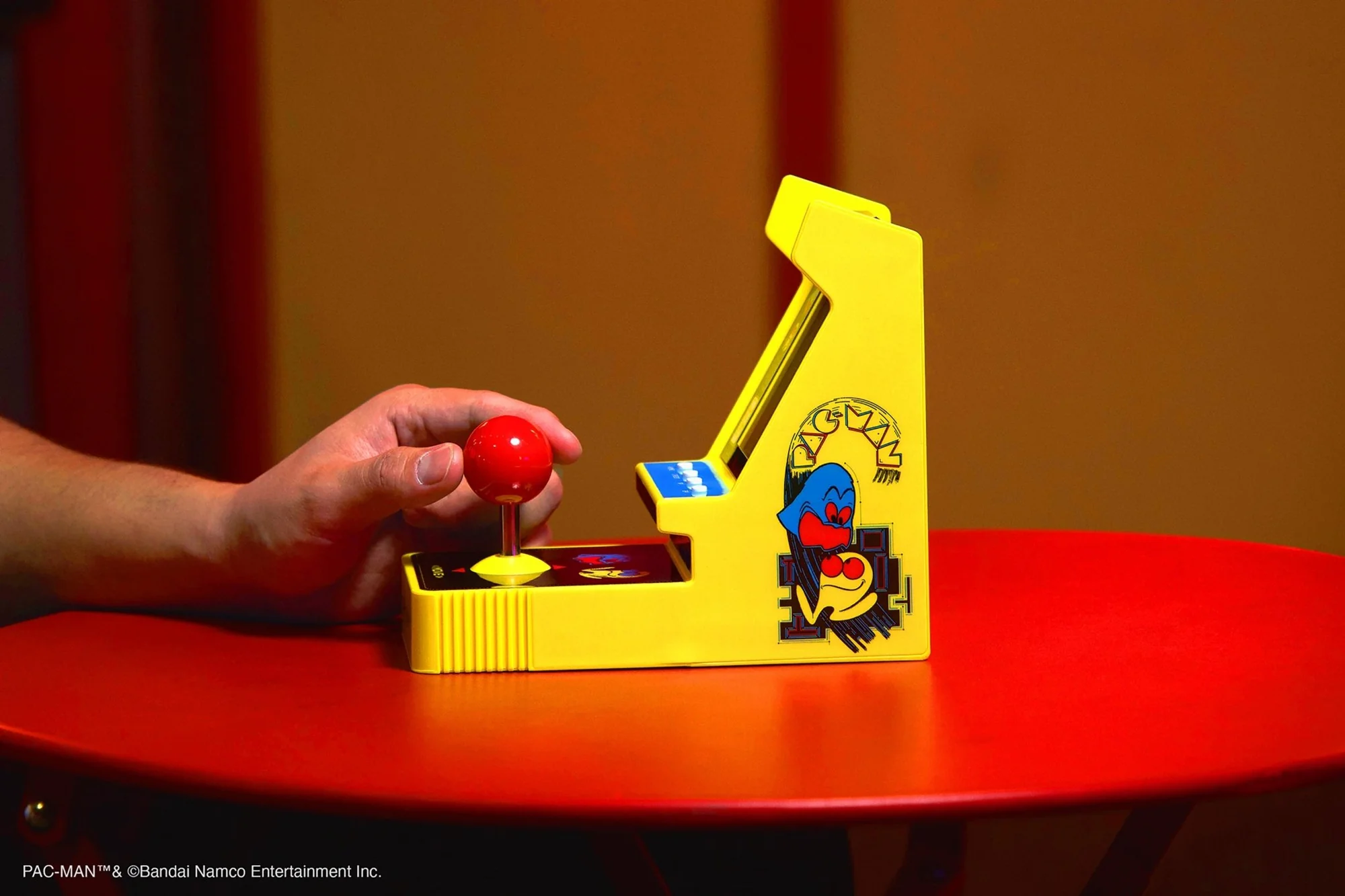 Une main tient un joystick rouge pour une borne d'arcade Pac-Man jaune. Le jeu est posé sur une table rouge.