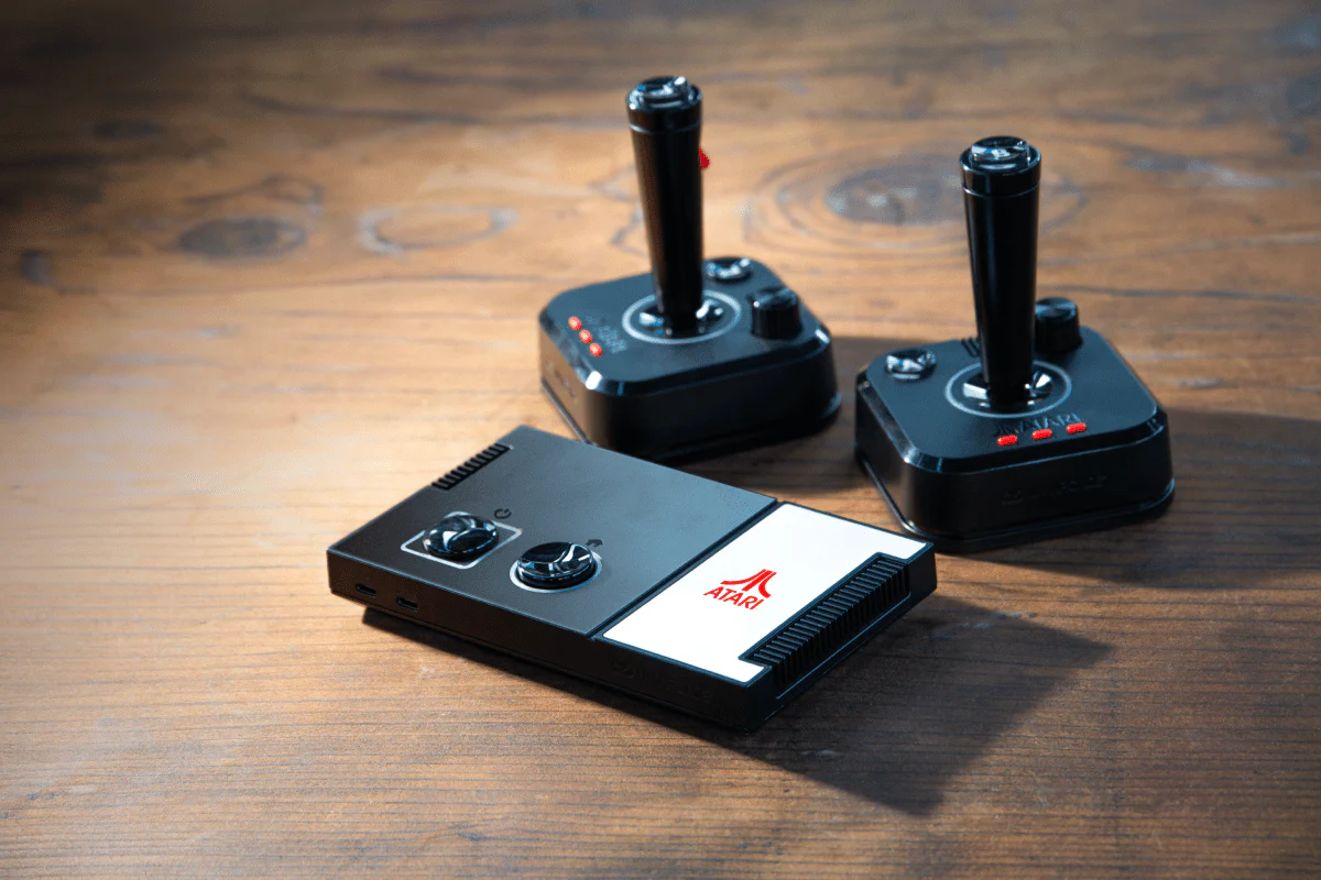 Trois manettes de jeu Atari classiques sur une surface en bois. Deux joysticks et une console, tous en noir et blanc.