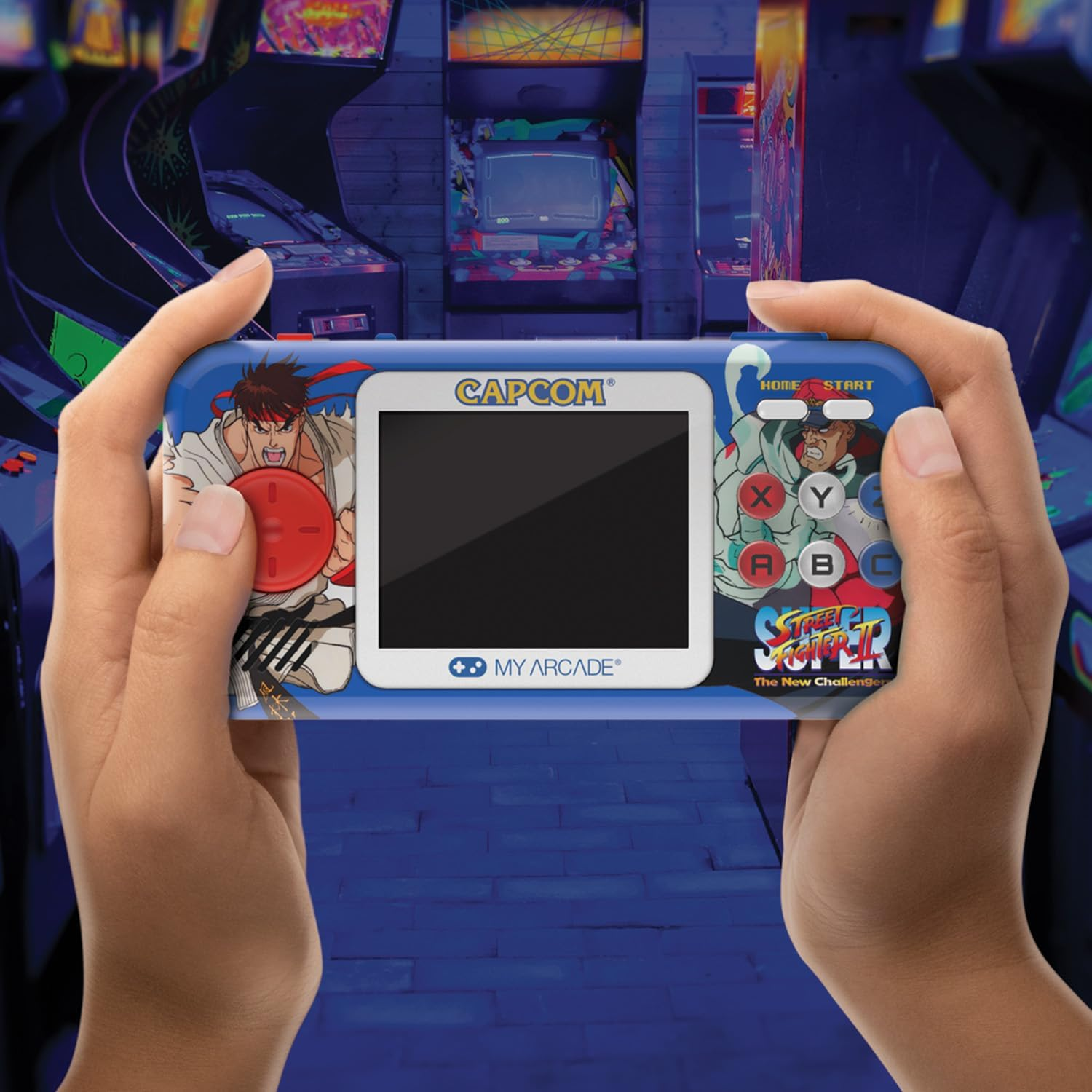 Des mains tiennent une console de jeu portable bleue My Arcade Street Fighter II. L'écran est noir. Des bornes d'arcade sont en arrière-plan.