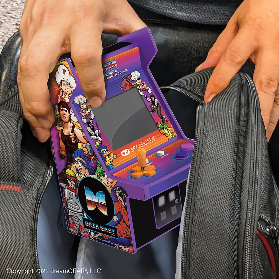 Mains tenant un jeu Data East My Arcade. Borne d'arcade violette avec joystick, boutons et écran, placée dans un sac noir.