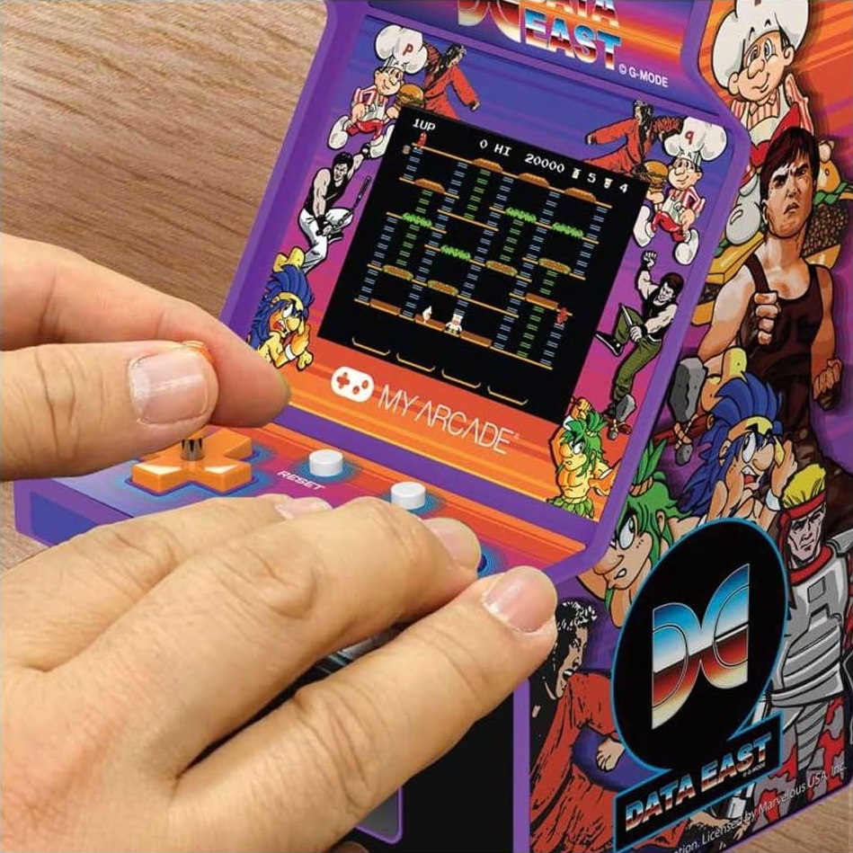 Mains jouant à un jeu d'arcade rétro. La console a un écran, un joystick et des boutons. Des graphismes colorés entourent l'écran.