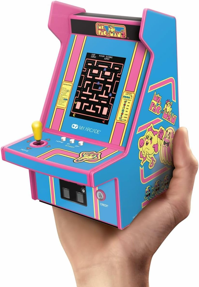Une main tient une mini-borne d'arcade Ms. Pac-Man. Elle est bleue et rose, avec un écran affichant le jeu. Les personnages de Ms. Pac-Man sont sur le côté.