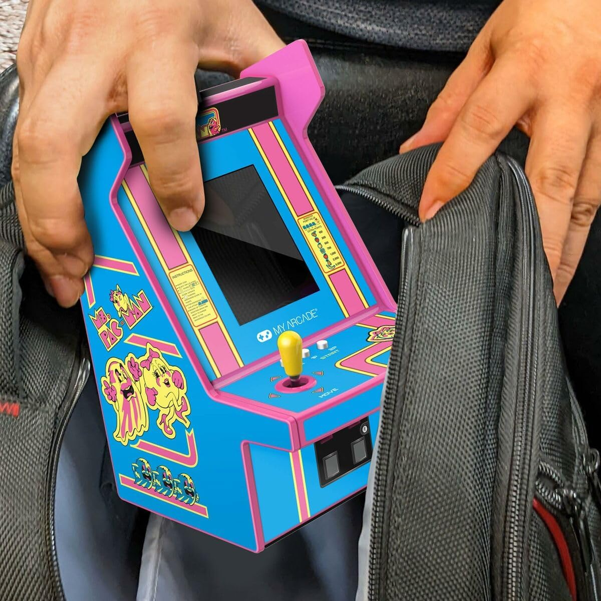 Mains tenant une mini borne d'arcade Pac-Man, bleue avec des bordures roses, la plaçant dans un sac gris. Joystick jaune et écran visibles.