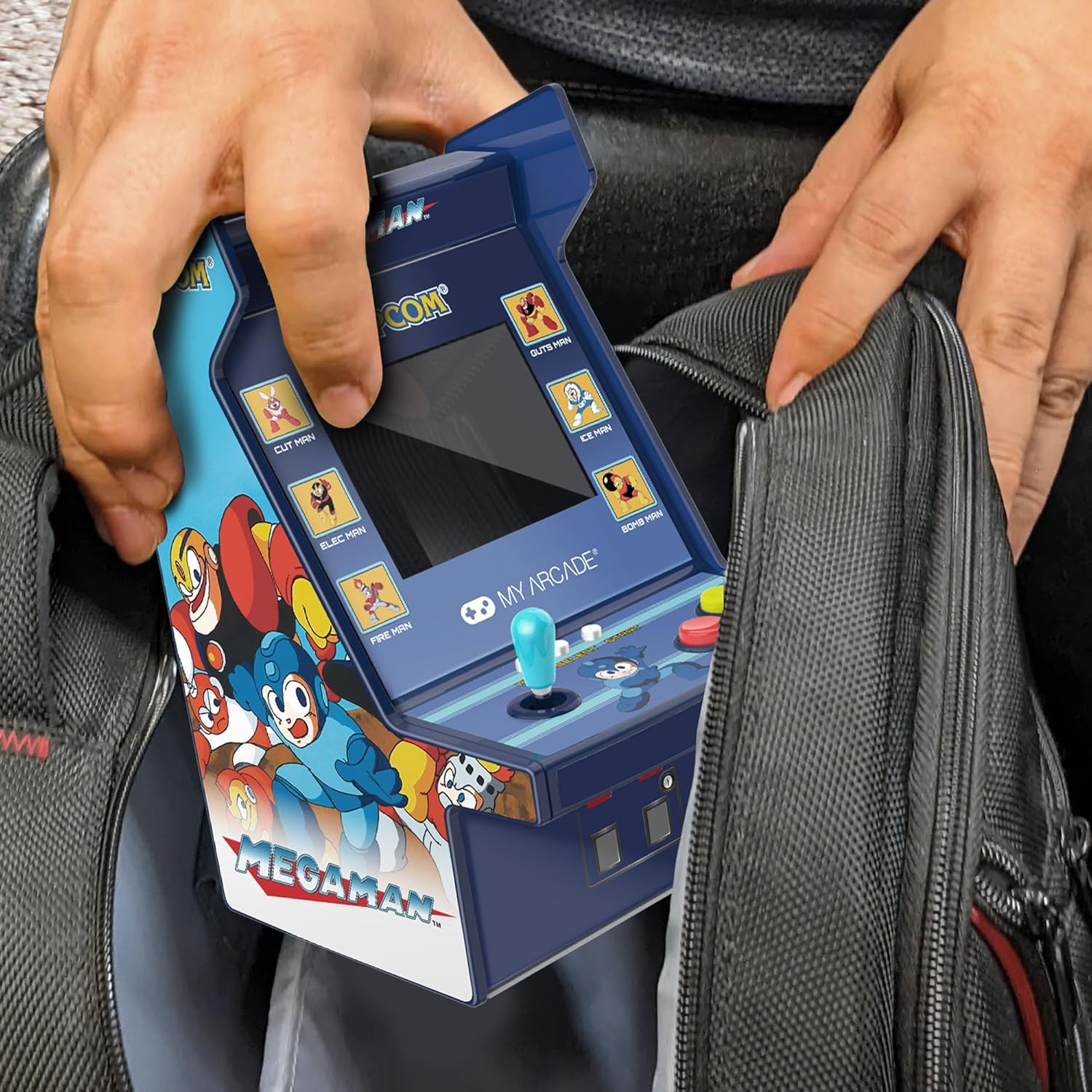 Des mains tiennent une borne d'arcade bleue Megaman, l'insérant dans un sac noir. Le jeu comporte des boutons et un joystick.