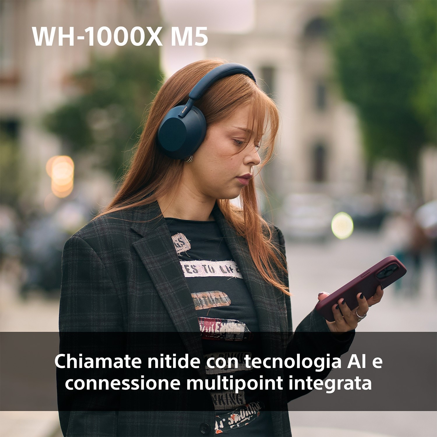 Donna con cuffie blu, tiene un telefono. Indossa un blazer nero ed è fuori. Il testo in alto dice WH-1000X M5.