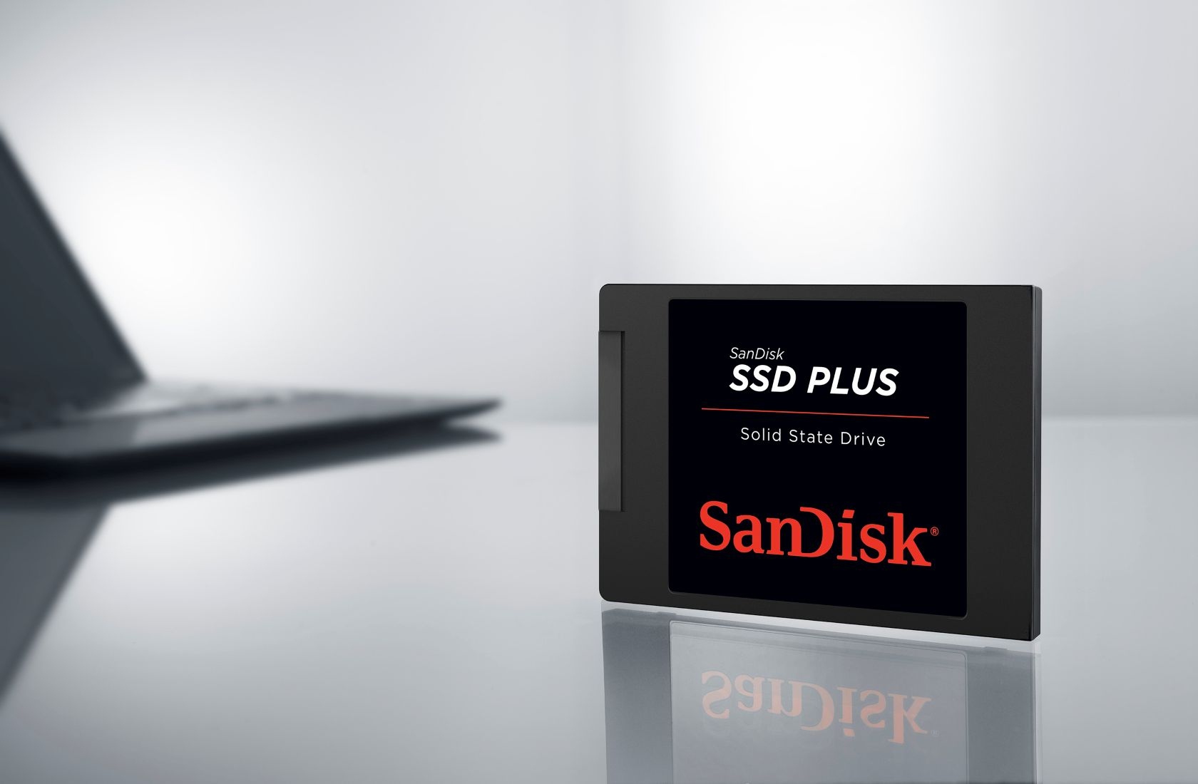 SSD Plus de SanDisk. SSD noire avec logo rouge et texte sur fond blanc. Ordinateur portable flou en arrière-plan.