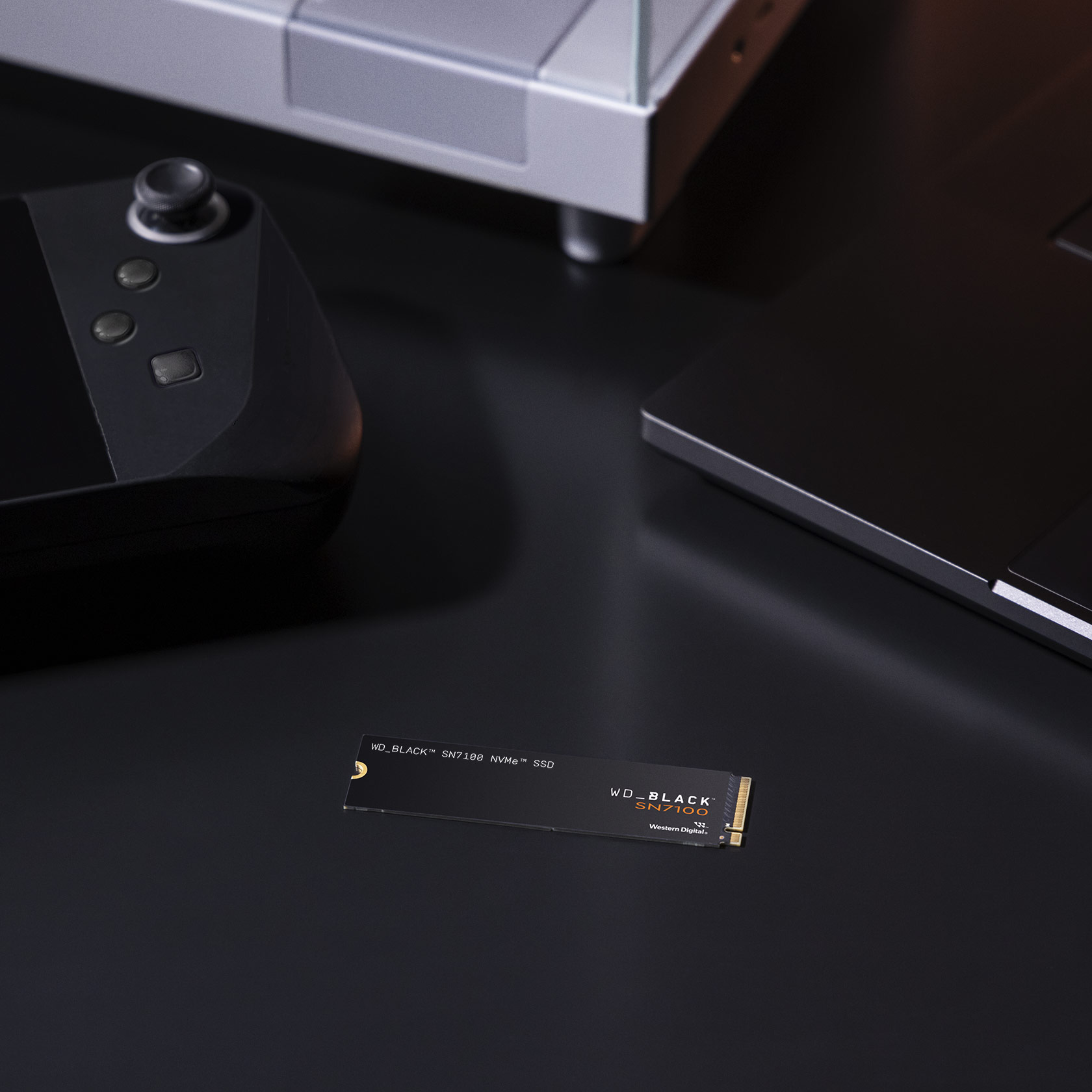 Schwarze SSD, Controller und Gamecontroller auf dunkler Oberfläche. Im Hintergrund ein Laptop und ein weißer Ständer.