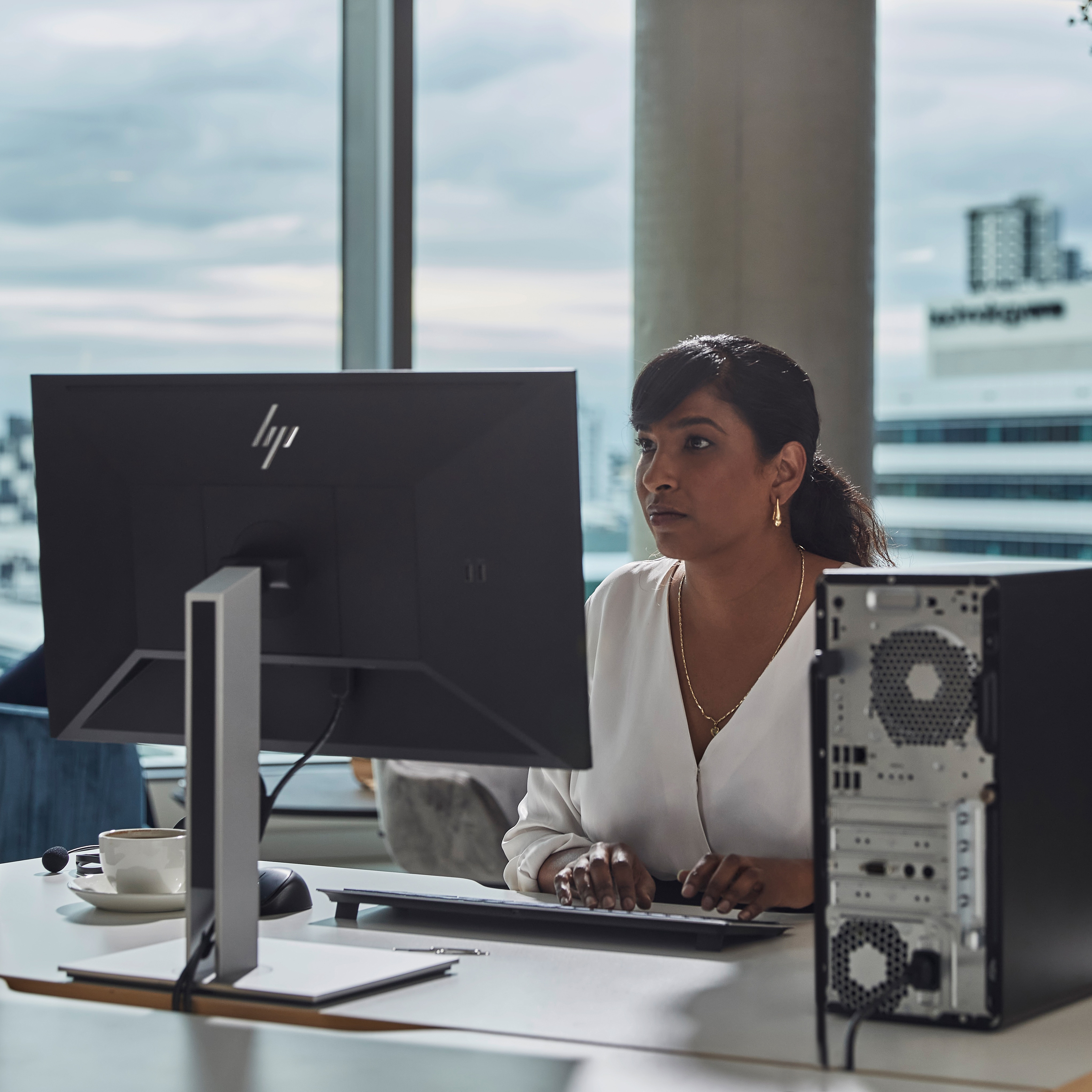 Eine Frau tippt an einem Schreibtisch auf einer Tastatur und blickt auf einen Computerbildschirm. Eine Tasse Kaffee und ein Computer sind ebenfalls auf dem Schreibtisch.