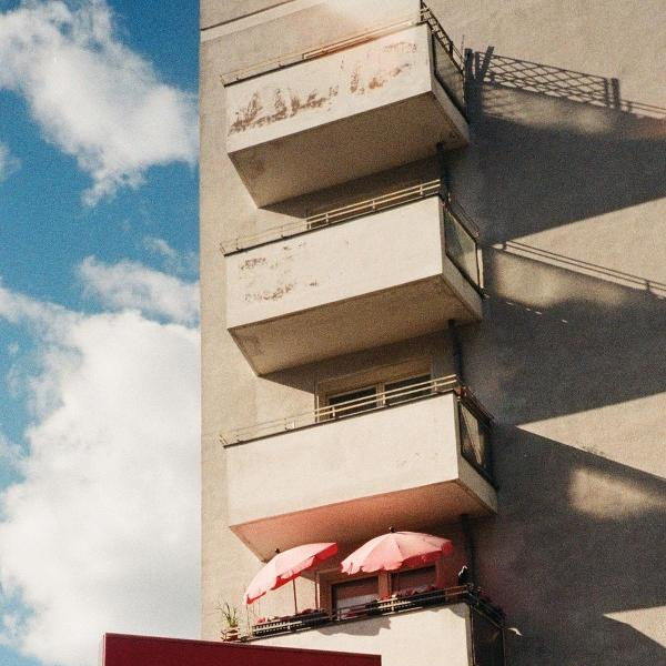 Ein Gebäude mit Balkonen und rosa Sonnenschirmen. Der Himmel ist blau mit Wolken. Schatten fallen auf das Gebäude.