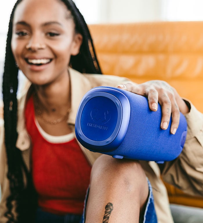 Une femme sourit, tenant un haut-parleur bleu. Elle porte un haut rouge, une veste beige et a un tatouage sur la jambe. Un canapé jaune est en arrière-plan.